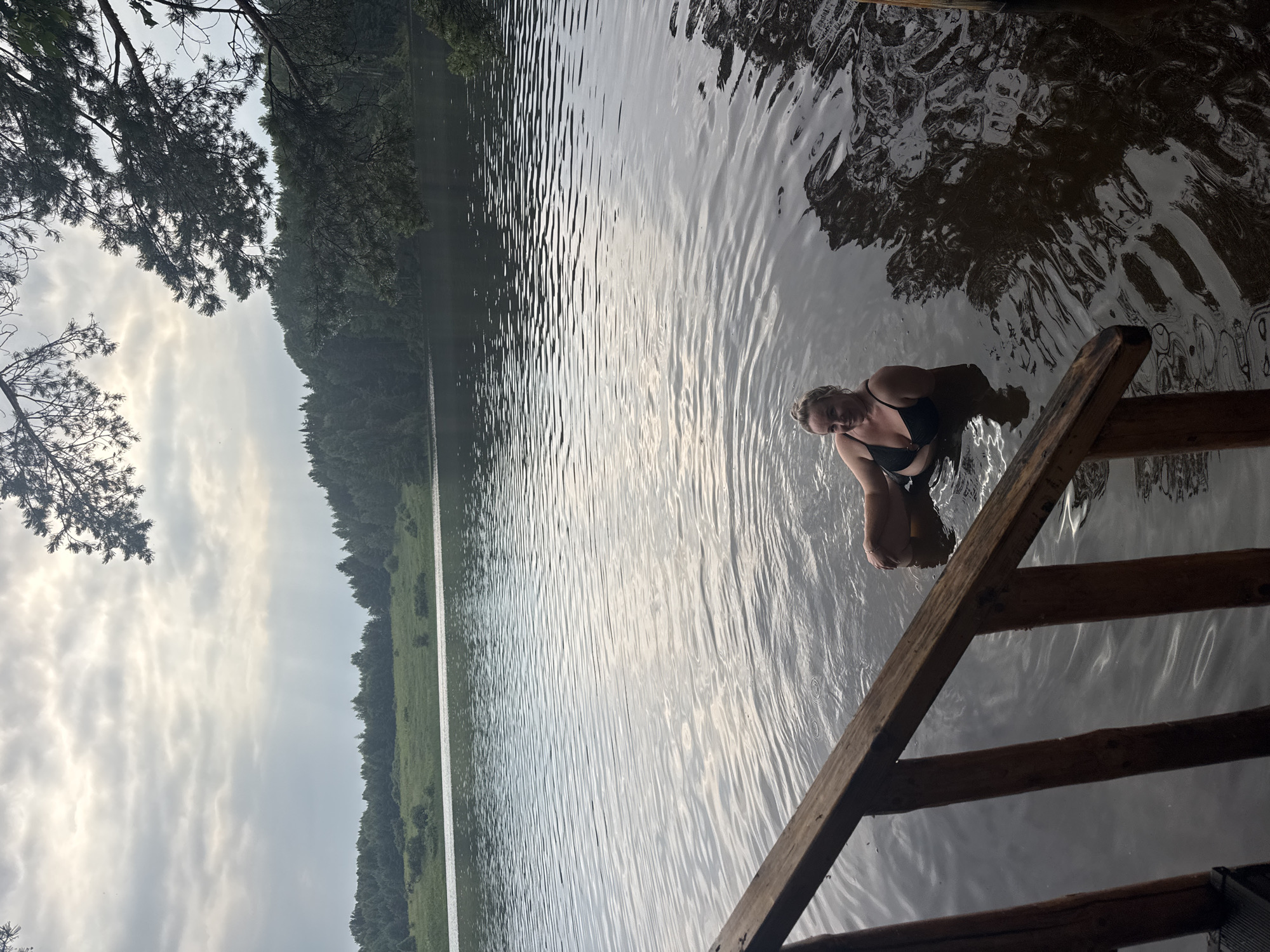 A person, a woman with blonde hair, is partially submerged in a body of water, sitting on a wooden ladder that extends into the water. She is wearing a black bikini. The water is calm with ripples reflecting the overcast sky. The background shows a lush green forest lining the opposite bank of the lake, with trees extending to the horizon. The sky is cloudy, suggesting a mild, possibly late afternoon or early morning, with diffused sunlight. The scene is set in a natural outdoor environment, likely a lake or large pond in Pozdyshki, Russia, indicated by the presence of trees and the calm water. The overall mood is tranquil and relaxed, with the woman appearing comfortable in the water. There is no visible text in the image.