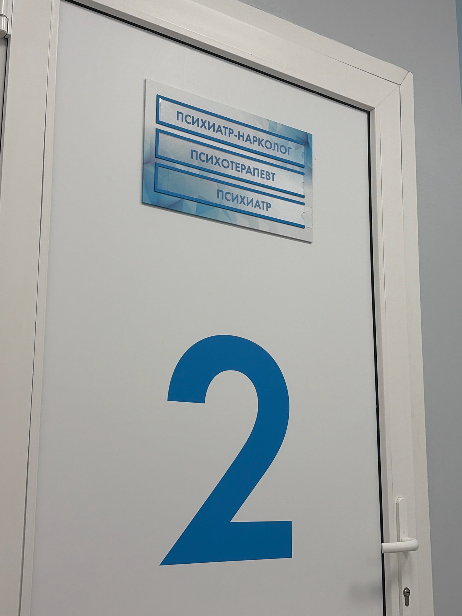 The image shows the interior of a door with a sign in Russian indicating various medical specialties. The sign has three sections, the top one reads "PSYCHIATRIST-NARCOLOGIST", the middle one reads "PSYCHOTHERAPIST", and the bottom one reads "PSYCHIATRIST". Below the sign, a large blue number "2" is painted on the door. The door has a white frame and a white handle with a keyhole. The scene is likely in a clinic or hospital in Arkhangelsk, Russia. The lighting suggests an indoor environment. There are no people visible in the image.