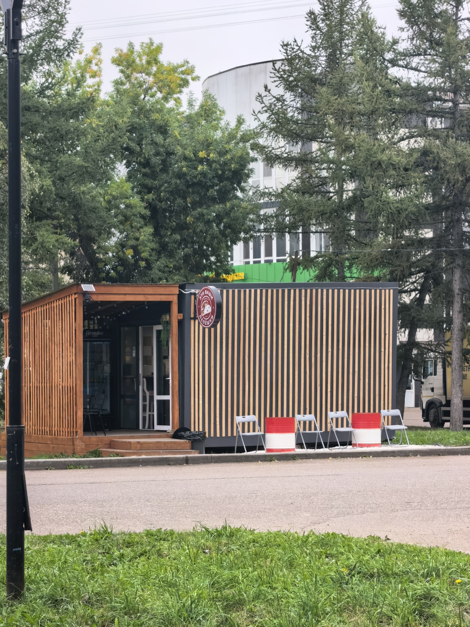 The image depicts a small, modern-style building, likely a cafe or food stall, with a wooden slatted exterior on one side and dark vertical wood slats on the other. The building has a wooden deck with steps leading up to an open entrance. Inside, a refrigerator and chairs are visible. A circular sign with text and an image is affixed to the dark-slatted side. In front of the building, there are four folding chairs and two cylindrical objects painted red and white, arranged along the edge of a paved area. Behind the building, trees and a multi-story building with windows are visible. A truck is parked to the right of the structure. The scene appears to be an outdoor commercial space in an urban setting. The location is identified as Sterlitamak, Russia.