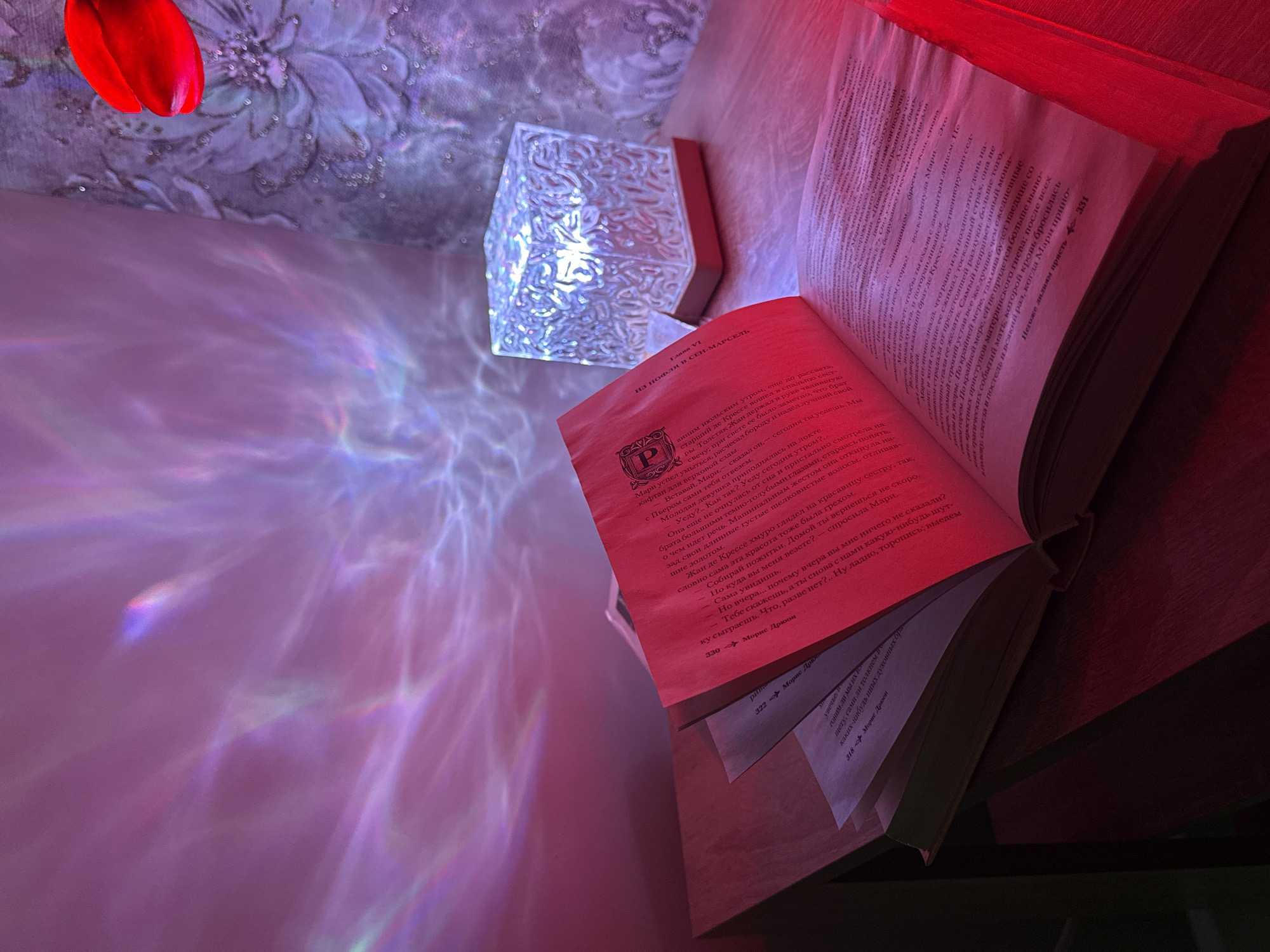 The image captures an open book resting on a wooden surface, illuminated by ambient light. The book has a red spine/cover and displays text in Russian. In the upper left corner, a bright red, possibly decorative, object hangs against a textured, floral-patterned background. To the right of this, a translucent, faceted object emits a cool, white light, casting colorful, refracted light patterns onto the surfaces below and to the left. The overall scene is bathed in a warm, reddish-pink glow, likely from another unseen light source or the red object in the upper left, creating a cozy and atmospheric setting, suggestive of evening or nighttime reading.
The book is open to pages featuring Russian text. Visible page numbers include "330", "322", "318", and "331". The author's name, "Морис Дрюон" (Maurice Druon), is clearly visible alongside the page numbers, indicating the book is by him. The chapter title "Глава VI НА НОФЛИ В СЕН-МАРСЕЛЬ" (Chapter VI IN HOFFLY IN SAINT-MARCEL) is displayed at the top of one page. A crest with the letter "P" is also seen on the page. The text describes characters named Mari and Jean, and mentions of "Крессе," "материнского гнева," and "Молодая девушка" are decipherable through OCR.