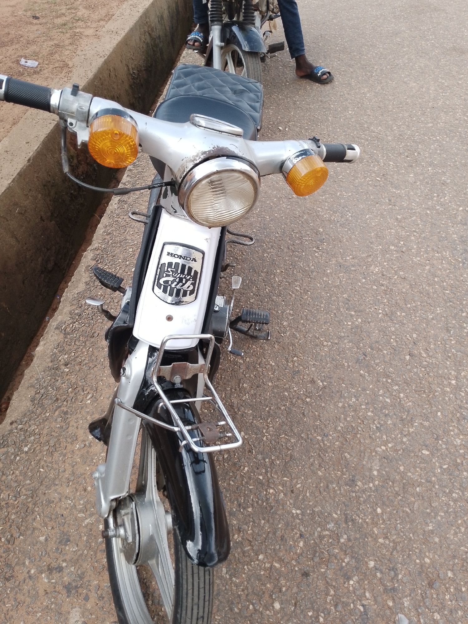 In Balewa, Nigeria, a close-up shot captures the front portion of a silver and black Honda Super Cub motorcycle. The handlebars, adorned with amber turn signals, are prominently featured. A black quilted seat is visible behind the handlebars, and the motorcycle's front fender is black. A chrome luggage rack is mounted above the front fender. The motorcycle is parked on an asphalt road, with a concrete drainage ditch running alongside it. A person's legs, clad in blue jeans and wearing black flip-flops, are visible in the upper right background, suggesting they are either the rider or a passenger of another motorcycle. The scene is illuminated by natural daylight.
