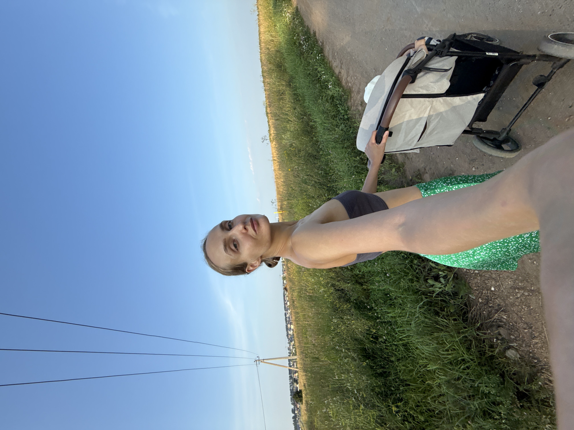 A woman is captured from the chest up in a selfie, walking along a dirt path in a rural area. She is wearing a dark gray bandeau top and a green skirt with a white floral pattern. Her blonde hair is pulled back in a ponytail. She is holding the handle of a light beige baby stroller with dark brown accents. The stroller has black wheels. In the background, there is a vast field of green grass and yellowing plants. A dirt road runs alongside the field. In the distance, a power line with three wires stretches across the frame, supported by a utility pole. Beyond the field, a cluster of houses with rooftops is visible, suggesting a nearby village or town. The sky is a clear, bright blue. The scene appears to be set in the village of Podshivalovo, Russia, given the context of the prompt.