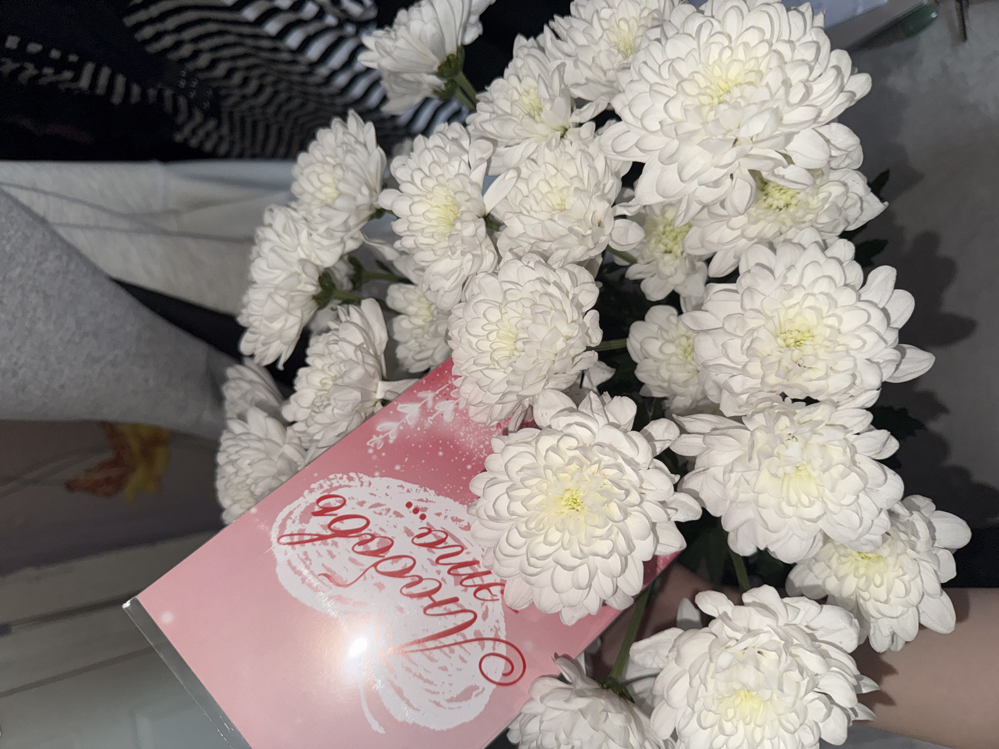 A medium close-up shot depicts a bouquet of white chrysanthemums and a pink card with Russian script, placed indoors. The flowers dominate the right side of the frame, their dense, layered petals creating a soft texture. A pink greeting card is nestled among the flowers, with a cursive red inscription in Russian, possibly spelling "Любимой Анне" (Beloved Anna). The card has a white, lace-like design. In the background, a striped fabric and a blurred surface are visible. The photograph does not explicitly state the location; therefore, I cannot confirm it is in Perm, Russia.