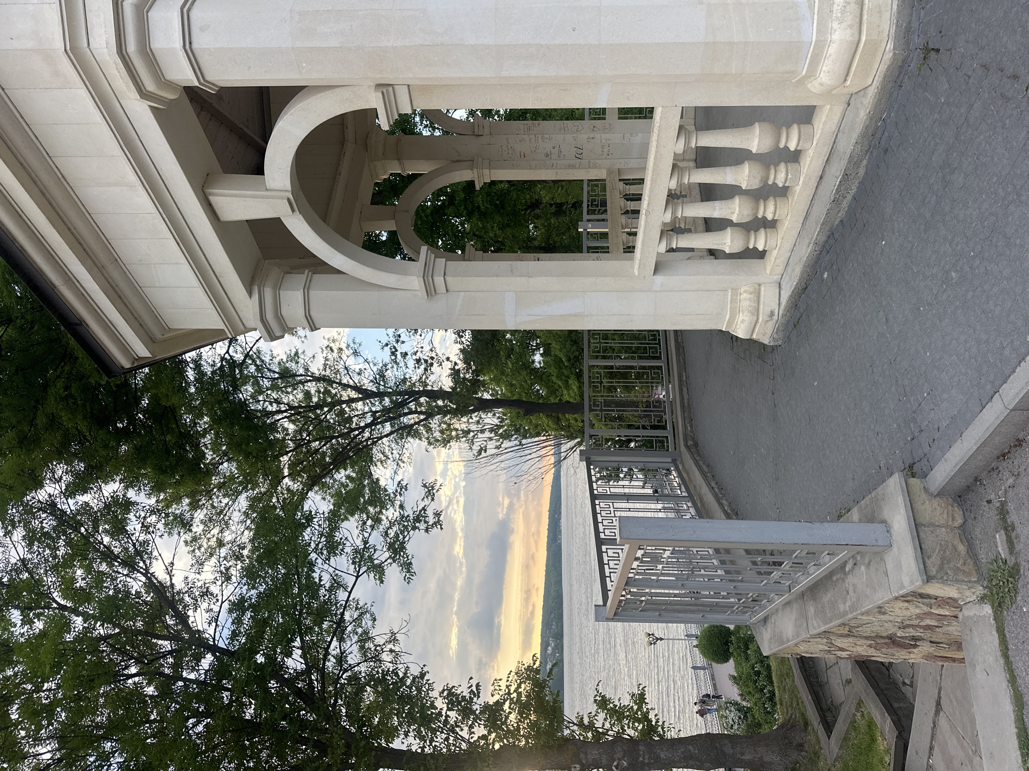 The image depicts a gazebo-like structure and a metal railing overlooking a body of water, likely the Ternopil Pond in Ternopil, Ukraine. The gazebo is constructed from light-colored stone with architectural details like arches, columns, and a balustrade. Graffiti is visible on the interior columns of the gazebo. A paved walkway is adjacent to the gazebo. Beyond the railing, trees frame a view of the pond, reflecting sunlight and clouds. In the distance, the opposite shore is visible, and small figures of people can be discerned near the water's edge. Vegetation, including trees and grass, is present on the near side of the pond.