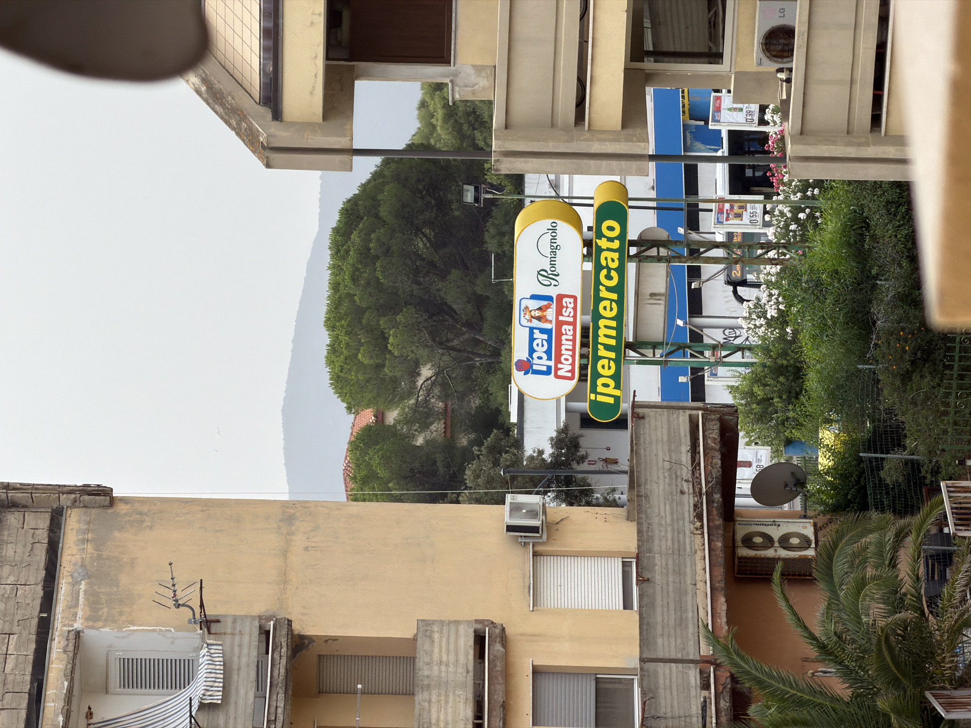 The image captures a view of buildings and vegetation in the city of Olbia, Italy, likely on an overcast day. In the foreground, a building with a yellow facade is visible, featuring balconies with concrete railings and shuttered windows. An air conditioning unit is mounted on the exterior wall. To the right, another building with a similar yellow facade and a brown-tiled roof section is present. A prominent sign with yellow and white lettering, advertising "ipermercato" (supermarket), is suspended between the buildings. Various trees, including palm trees and dense green foliage, are scattered throughout the scene. In the background, rolling hills are faintly visible under the hazy sky. A street lamp and other utility poles are also present. The overall impression is that of an urban environment with significant greenery.