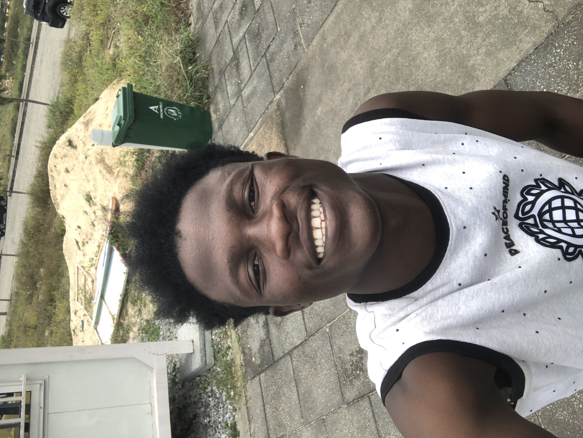 A medium shot captures a young Black man in a sleeveless white shirt with black trim and a black polka dot pattern, standing outdoors. He is smiling and looking directly at the camera. His dark hair is short and curly. The background includes a paved walkway, grass, a pile of sand, a white structure, and a green trash bin. A black SUV is visible in the distance. The scene appears to be in Ikoyi, Nigeria.