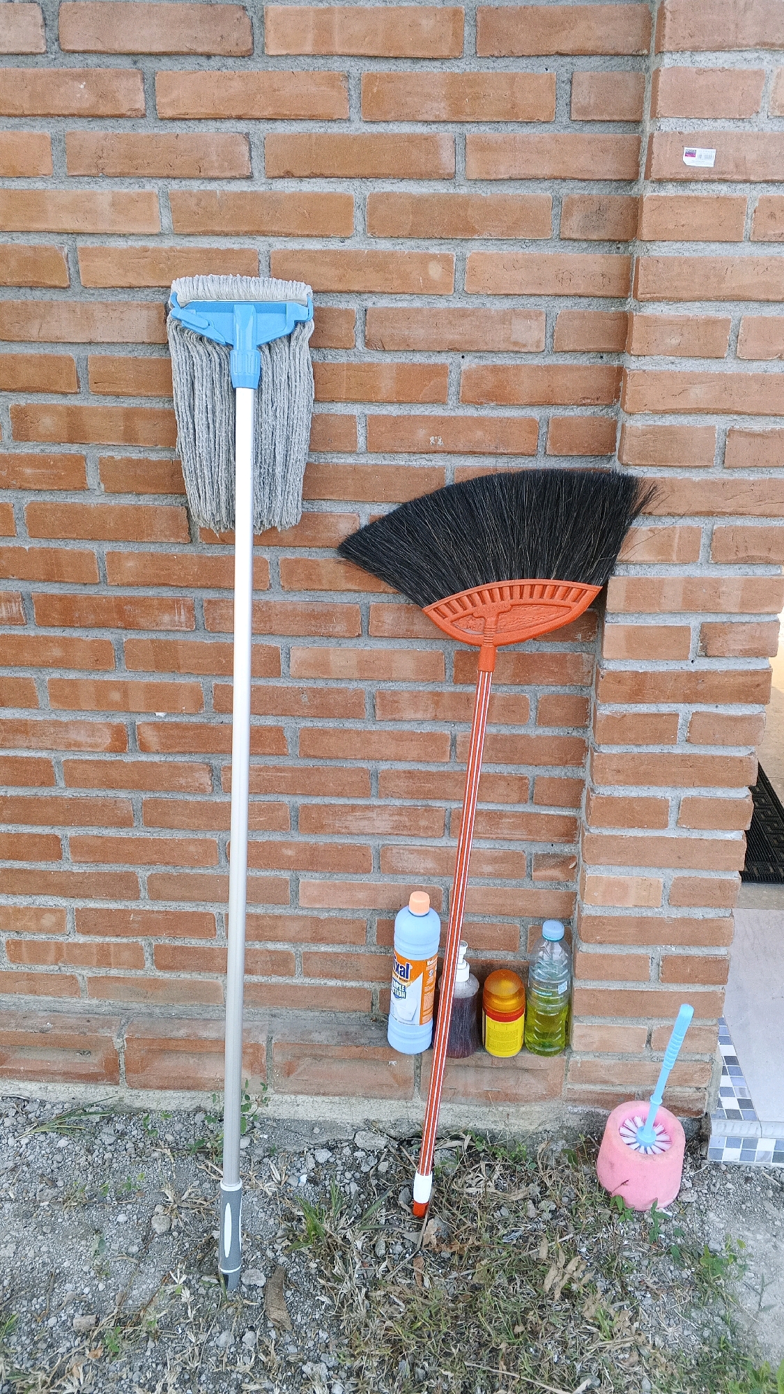 This image shows a collection of cleaning supplies leaning against a brick wall. The scene is outdoors, with dry grass and gravel on the ground. The cleaning supplies include a mop with a blue head and a silver handle, an orange broom with black bristles, and several bottles of cleaning liquids. A pink toilet brush with a blue handle is also visible. The brick wall is made of reddish-brown bricks with gray mortar. The lighting suggests it is daytime, with shadows cast by the cleaning supplies. The location is identified as Minggir, Indonesia, though no specific landmarks or identifying features of the city are visible in the image itself. There are no people present in the image.