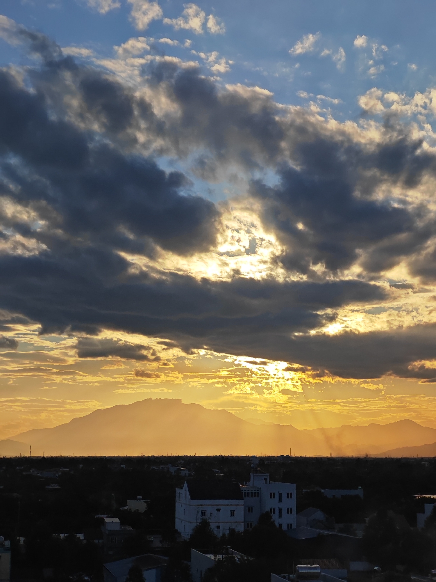 This image captures a dramatic sunset over Đà Nẵng, Vietnam, viewed from a high vantage point. The sky is a dynamic canvas of contrasting colors and textures, with a brilliant golden-orange glow dominating the lower half, illuminated by the setting sun peeking through dark, heavy clouds. The sun's rays pierce through gaps in the clouds, creating a dramatic, almost ethereal effect.
In the mid-ground, a long, silhouetted mountain range stretches across the horizon, softened by the hazy sunset light. Below the mountains, the urban landscape of Đà Nẵng is visible as a dark silhouette, with the outlines of buildings and trees discernible. A prominent white building with multiple windows stands out amongst the darker structures in the foreground. The overall atmosphere is one of natural beauty and the convergence of urban life with a powerful sky.