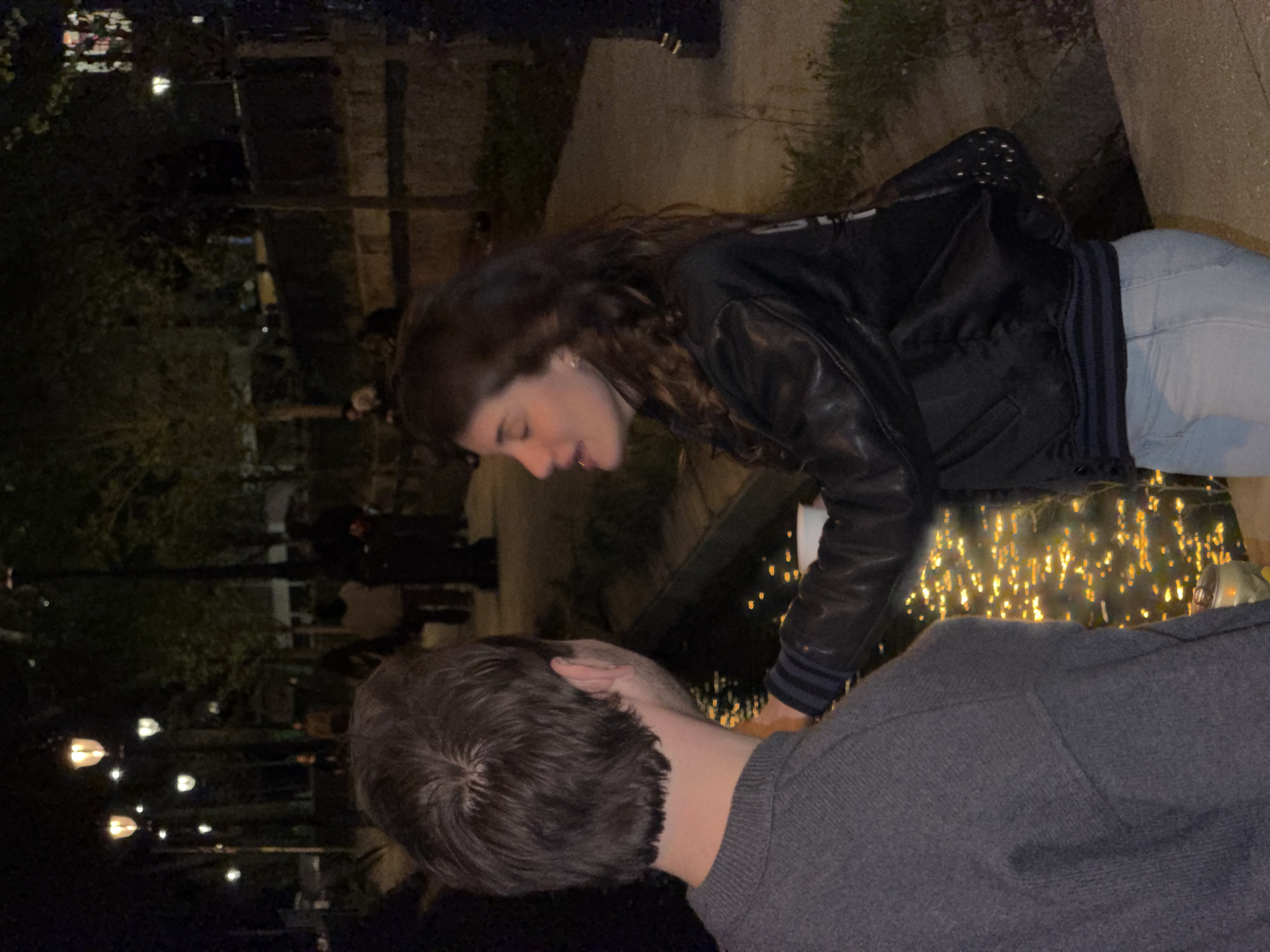 A couple is positioned in front of a body of water, possibly a canal or pond, reflecting numerous small lights at night. The woman has shoulder-length brown hair and wears a black leather jacket and light blue jeans. She faces down towards the man, potentially speaking to him or observing something in the water. The man has short, dark hair and is wearing a gray sweater. His back is mostly visible, with the back of his head turned upwards towards the woman. The background shows trees and the silhouettes of people walking. There are also lamps visible in the background. The scene appears to be in an urban setting at night, though specific Parisian landmarks are not definitively visible.