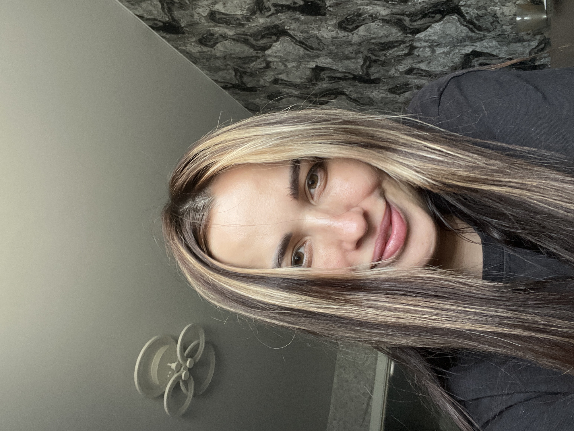 A close-up, slightly rotated, selfie depicts a young woman with brown hair and blonde highlights. She has fair skin, brown eyes, and a slight smile. She is wearing a dark-colored top. The background consists of a gray wall with a white decorative element and a wall with a textured, dark gray pattern. The location is Unknown, Unknown.