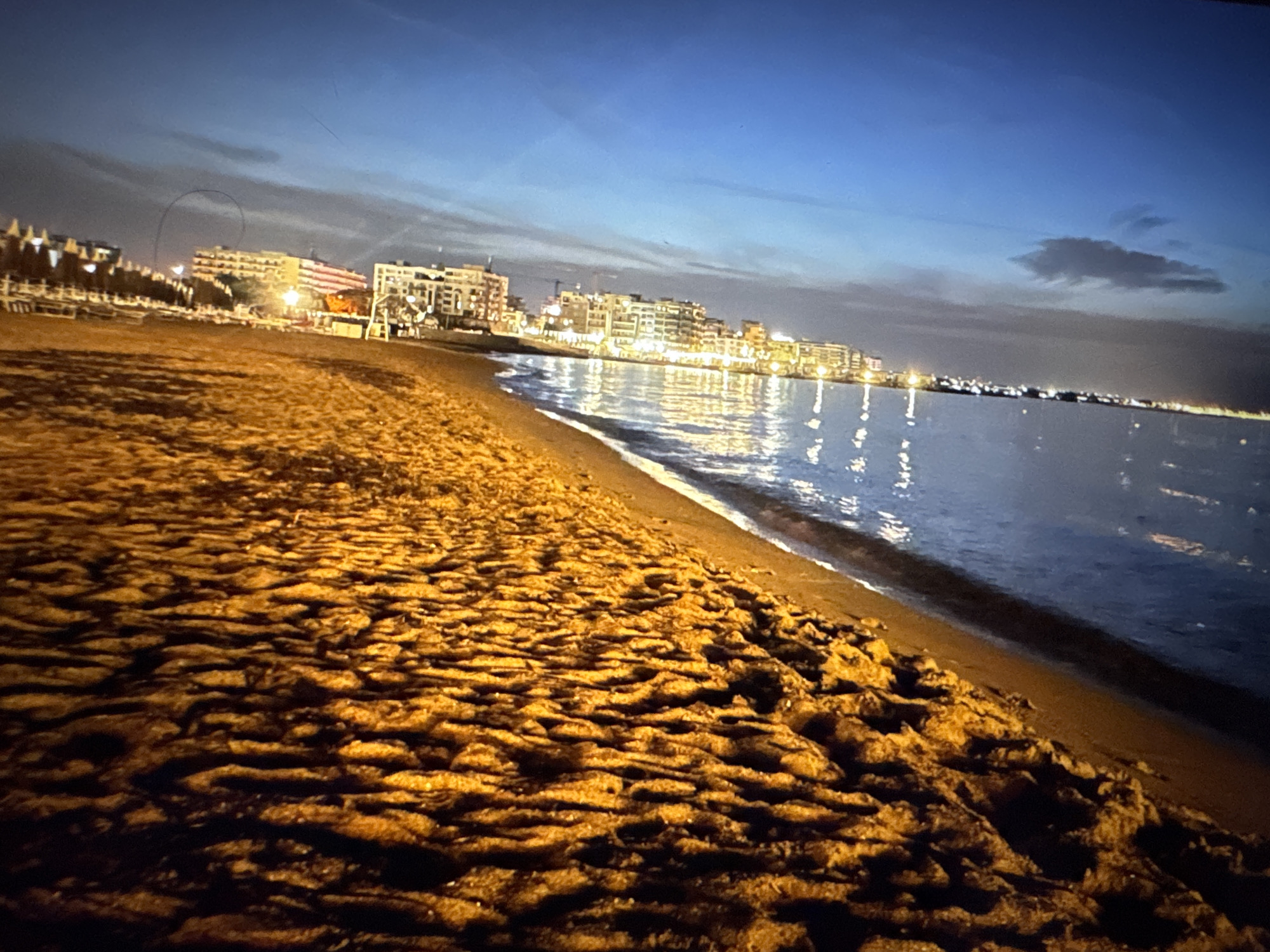 The image shows a beach at dusk or dawn, with a calm sea on the right and buildings illuminated by artificial lights in the distance. The sand in the foreground is textured with ripples and shadows, suggesting it has been disturbed by wind or water. The sky is a gradient of deep blue to lighter blue, with some clouds visible. The buildings appear to be hotels or apartments, lining a waterfront. The scene is devoid of people. The location is not Porrentruy, Switzerland, as Porrentruy is an inland town.