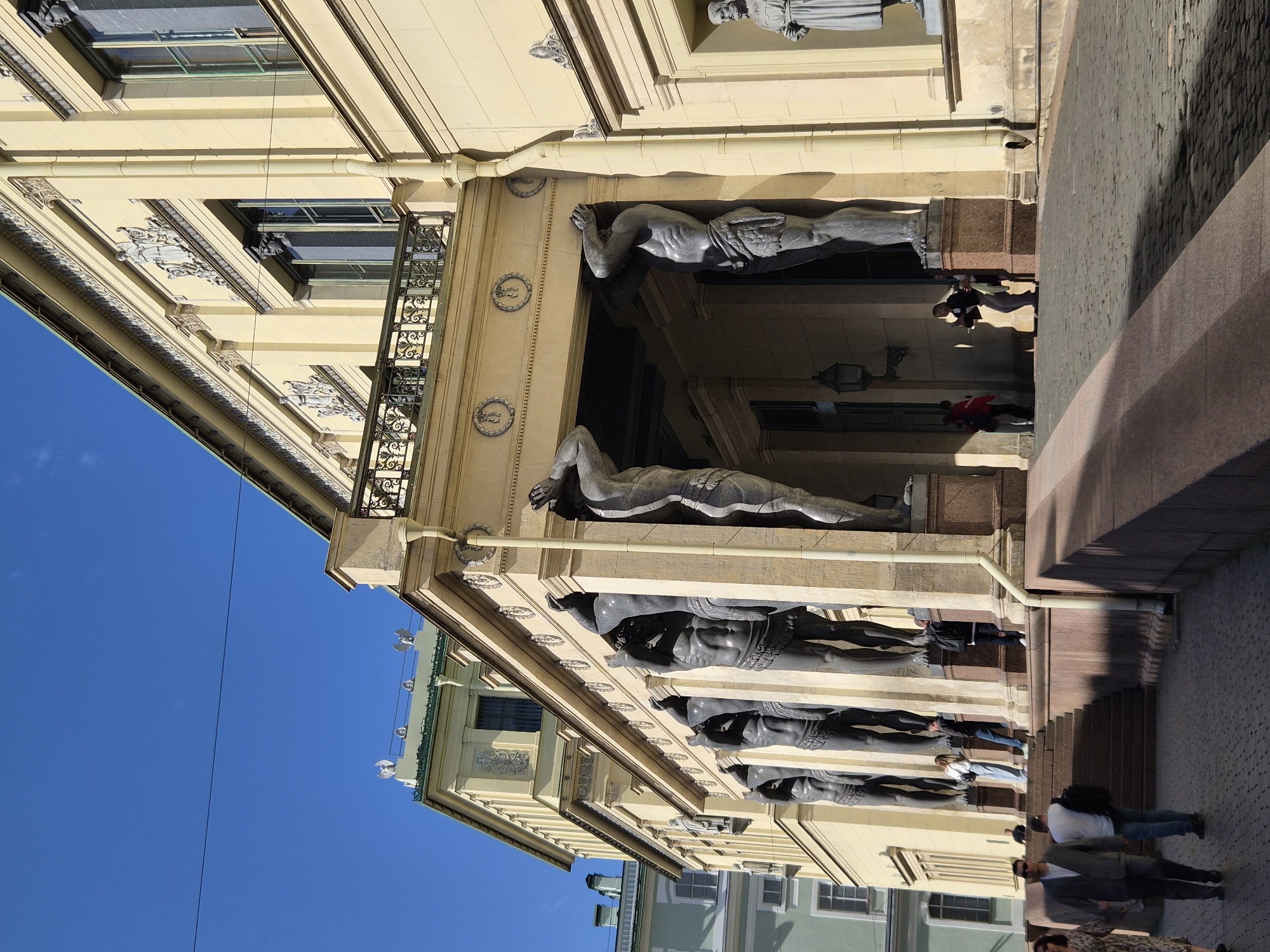 The image captures a side view of a grand building with a yellow facade, adorned with classical architectural elements. In the foreground, a series of colossal statues of men, likely Atlantes, support an ornate balcony. These statues are dark grey and appear to be made of stone. A wide, stone staircase leads up to the building's entrance, with several people ascending and descending.
To the left of the staircase, a cobblestone pathway is visible, with more pedestrians walking. The building's facade features large windows, decorative friezes, and ornate moldings. Above the balcony, a portion of the building's upper stories is visible, also in a pale yellow hue, with a green roofline in the distance. The sky above is a clear, bright blue. The overall scene suggests a sunny day in a location of significant architectural importance.
