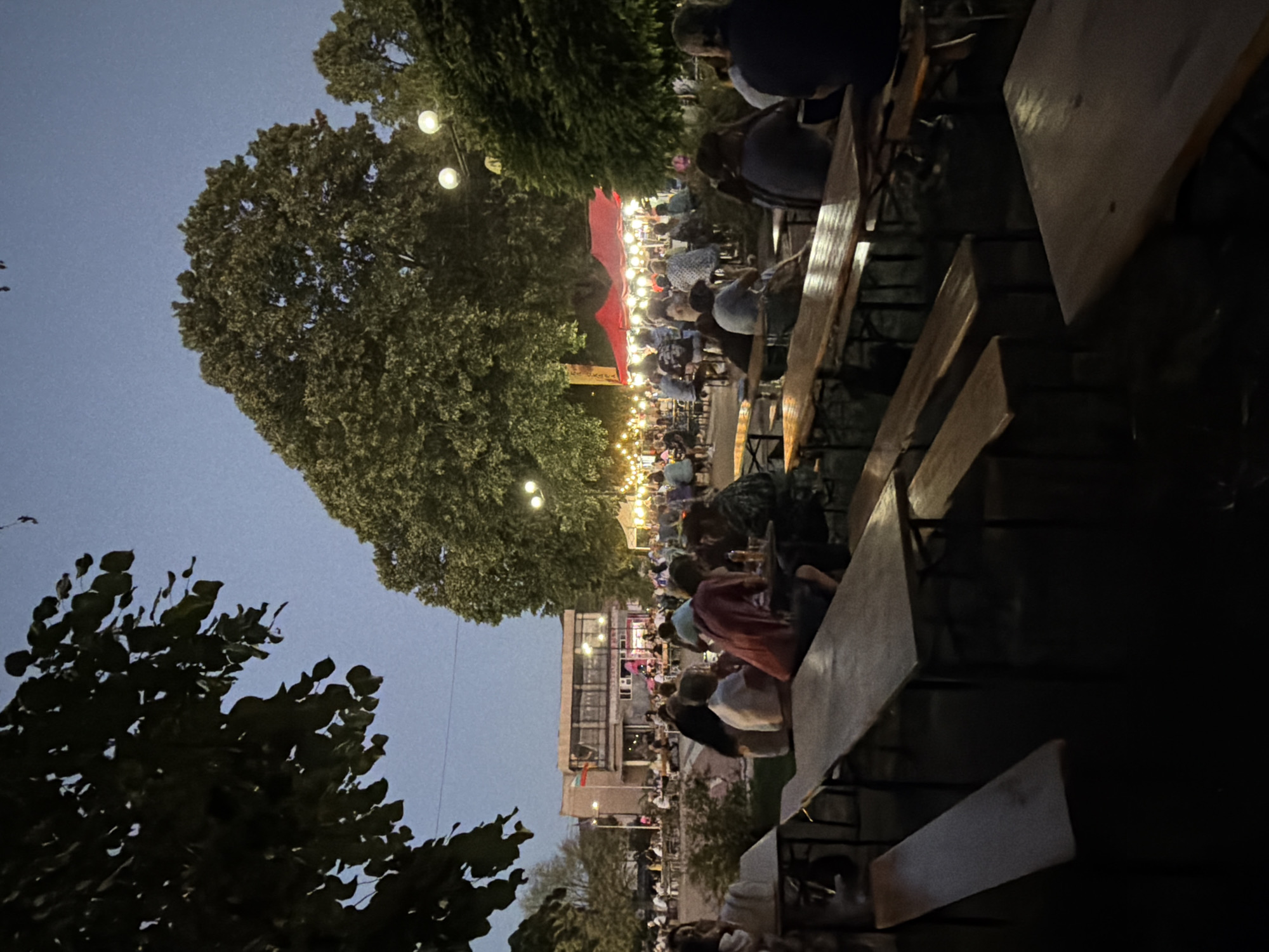 An outdoor evening gathering is depicted, likely a festival or market, in the city of Mursalevo, Bulgaria. The scene is illuminated by strings of festoon lights and a few street lamps, casting a warm glow on the assembled crowd. Large trees, silhouetted against the darkening sky, frame the upper left and right portions of the image. In the background, a building with large windows and flags is visible. Numerous wooden picnic tables and benches are arranged throughout the area, with people seated at many of them, engaged in conversation or enjoying food and drink. Some individuals are standing and mingling. Red tents, possibly stalls or food vendors, are set up further back in the scene. The overall atmosphere appears relaxed and social, with a sense of community evident amongst the attendees. The ground surface is not clearly discernible but appears to be a paved or earthen area.