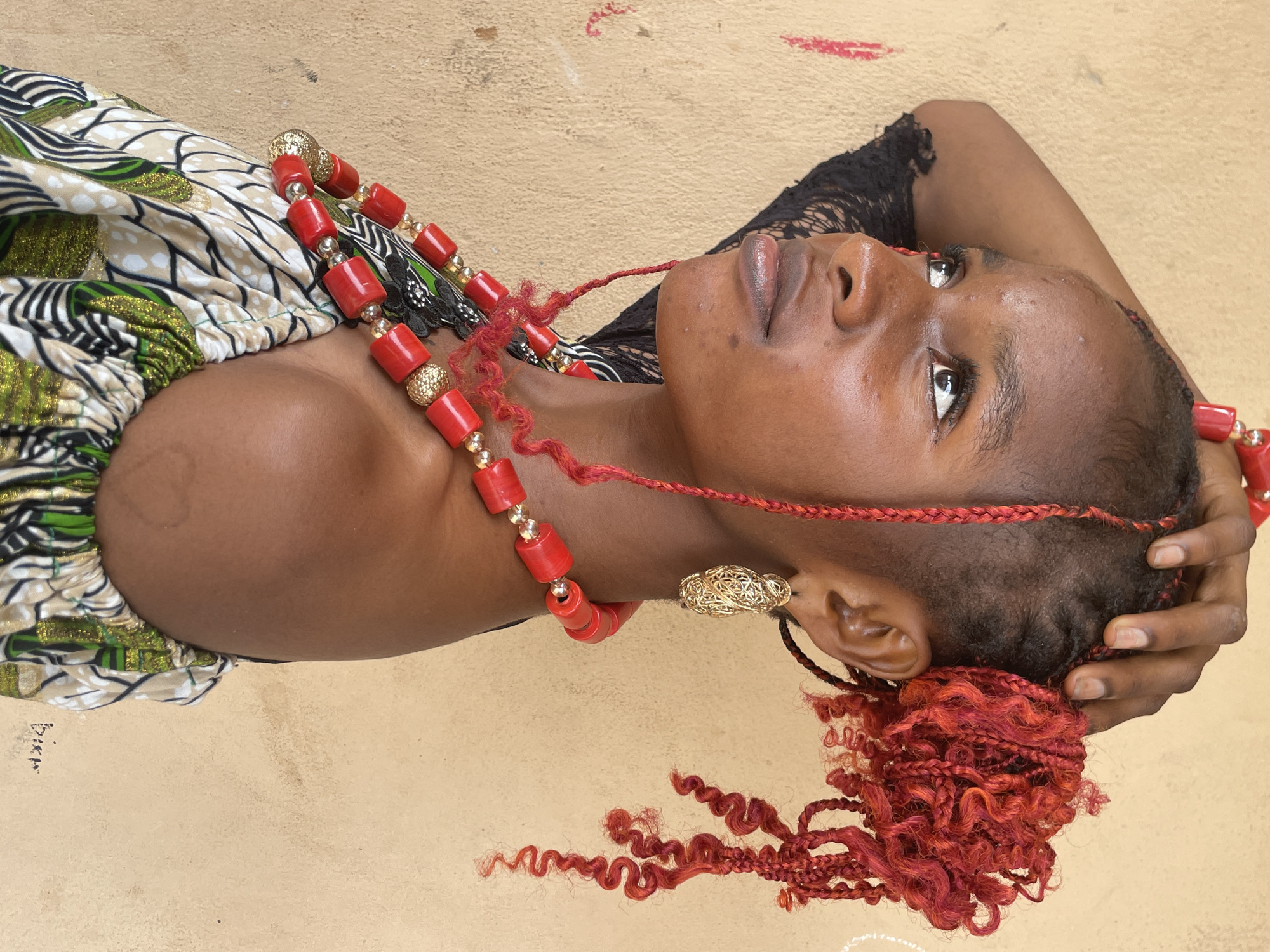 A close-up, high-angle shot captures a young Black woman lying down, looking upwards with a serene expression. Her head is turned to the left, showing her profile. She has dark skin and full lips. Her hair features red braids and curls, with a shaved or closely cropped section on the side. One arm is bent, with her hand resting behind her head, partially obscured.
She wears a prominent red and gold beaded necklace around her neck, and ornate golden filigree earrings are visible on both ears. Her attire includes a colorful, patterned garment, possibly made from Ankara fabric, with green, white, black, and gold designs. A black lace garment is visible near her shoulder and neck.
The background is a plain, light beige or sandy-colored surface, possibly a wall or ground. There are faint red smudges in the background above her head. The lighting is bright and even, suggesting daytime. In the bottom left corner, near her arm, the faint text "Bikin" (or similar script) is visible, appearing to be a signature or watermark. The subject's attire and features are consistent with the provided context of Badagry, Nigeria.