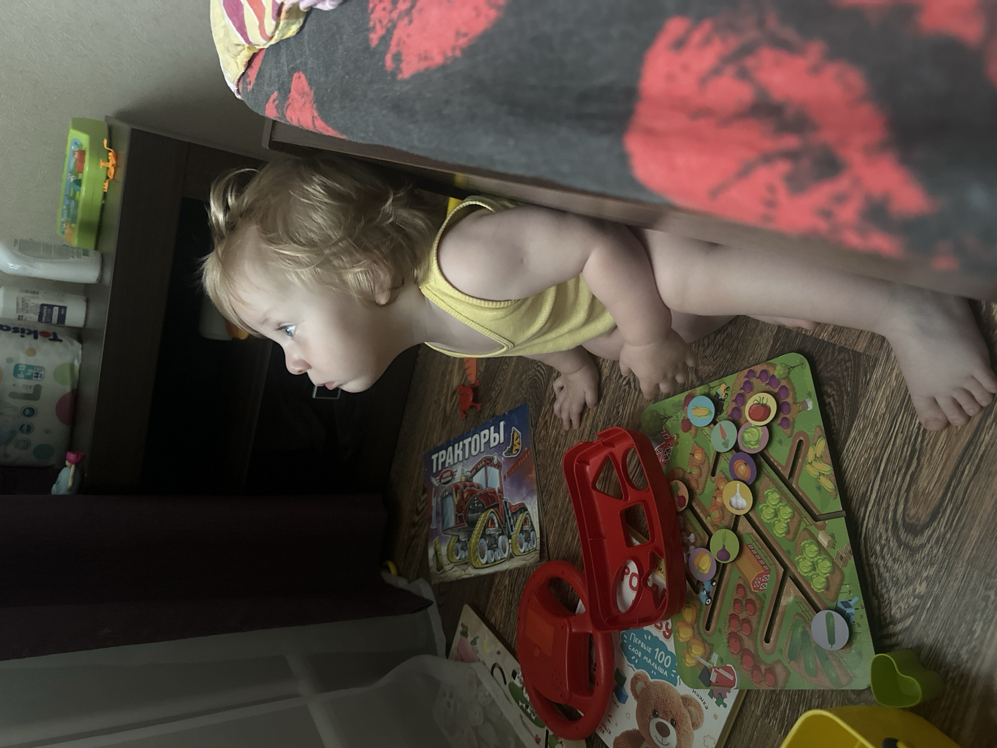 A young child with blonde hair is lying on the floor, looking to the left. The child is wearing a yellow tank top. The floor is made of wooden planks. Several toys are scattered around the child, including a red steering wheel toy, a green maze game with vegetable pieces, and a book with a tractor on the cover. The book has Russian text that reads "ТРАКТОРЫ" (Tractors). Next to the book are other children's books, one with a picture of a teddy bear and Russian text that says "ПЕРВЫЕ 100 СЛОВ МАЛЫША" (The First 100 Words of a Baby). In the background, there are shelves with some bottles and a green toy. A blanket with a red pattern is draped over a piece of furniture above the child. The scene appears to be indoors, possibly in a child's room or play area. The lighting suggests it is daytime.