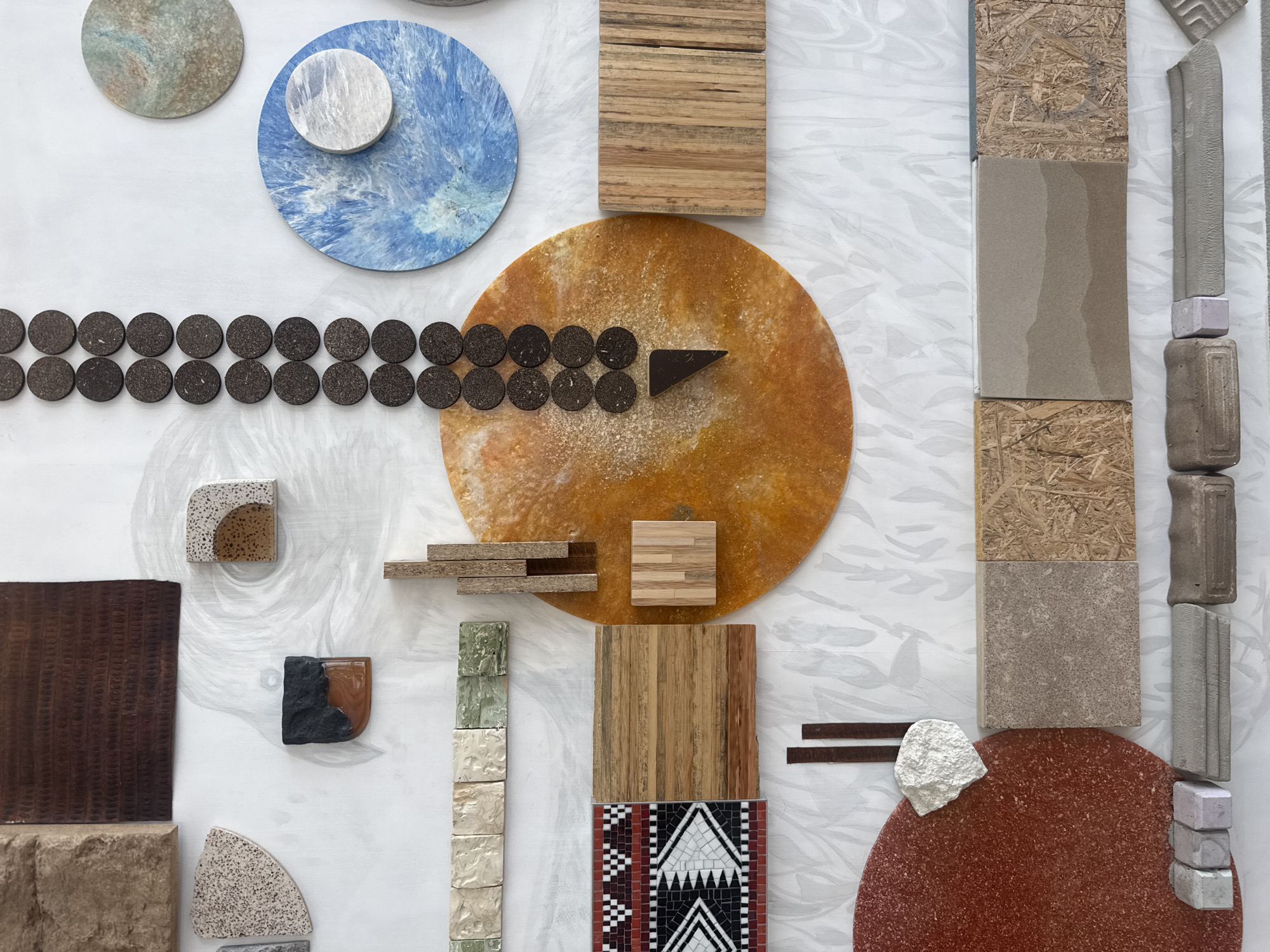The image displays a variety of material samples arranged on a white patterned background. At the top left are two circular tiles, one pale green/gray and the other a blue swirl with a smaller white circle on top. Below them is a horizontal line of dark brown circular objects, possibly cork. To the right is a large orange circular sample with a scattering of light-colored material, and a dark brown triangular piece resting on it.
Several small wooden blocks and strips are scattered between the left and center. A textured dark brown square is on the left. Moving down, there is a row of varying colored stone tiles, a square of wooden vertical strips, and a mosaic tile with red, white, and black geometric patterns.
On the right side, starting from the top, are samples of composite wood, beige stone, and a column of stacked concrete bricks. A couple of thin brown rectangular strips rest near a rough white stone, atop a red-brown circular tile. The bottom right finishes with light gray blocks arranged vertically.
There are no people visible in the image, and the objects appear to be material samples for a design or construction project. There is no direct information regarding the location being Dubai, United Arab Emirates, present in the image.