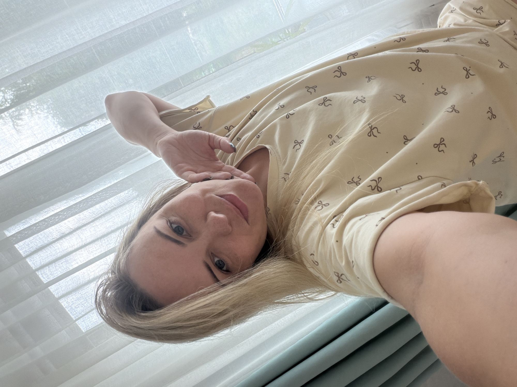 A woman with blonde hair and blue eyes is taking a selfie. She is wearing a light-colored pajama top with a bow pattern. Her left arm is bent with her hand resting on her cheek. She is positioned in front of sheer white curtains that are partially drawn over a window. A green curtain is visible on the left side of the frame. The scene suggests an indoor setting, likely a bedroom. There is no information to indicate the location is Votkinsk, Russia.