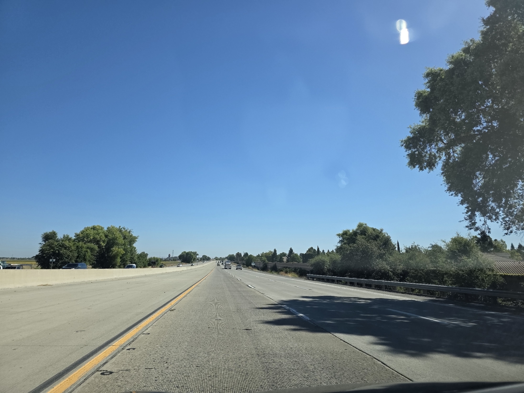 This media file captures a view from a vehicle driving on a highway in Sacramento, California. The scene is set on a sunny day with a clear blue sky, indicating good weather. The highway stretches into the distance, with multiple lanes of concrete pavement marked by yellow and white lines. Several vehicles, including cars and trucks, are visible on the road, suggesting moderate traffic.
To the left of the highway, a concrete barrier separates the lanes from an adjacent area with a few scattered vehicles and a line of green trees. To the right, a metal guardrail runs alongside the road, separating it from a residential area with houses and more lush greenery. A large tree with dark green foliage is prominently featured on the right side of the frame, with its branches extending towards the sky.
The time of day appears to be late morning or early afternoon, given the bright sunlight and the shadows cast on the road. There are no people clearly visible in the media file. No text is discernable in the image. The overall impression is one of a typical day of travel on a suburban or exurban highway.