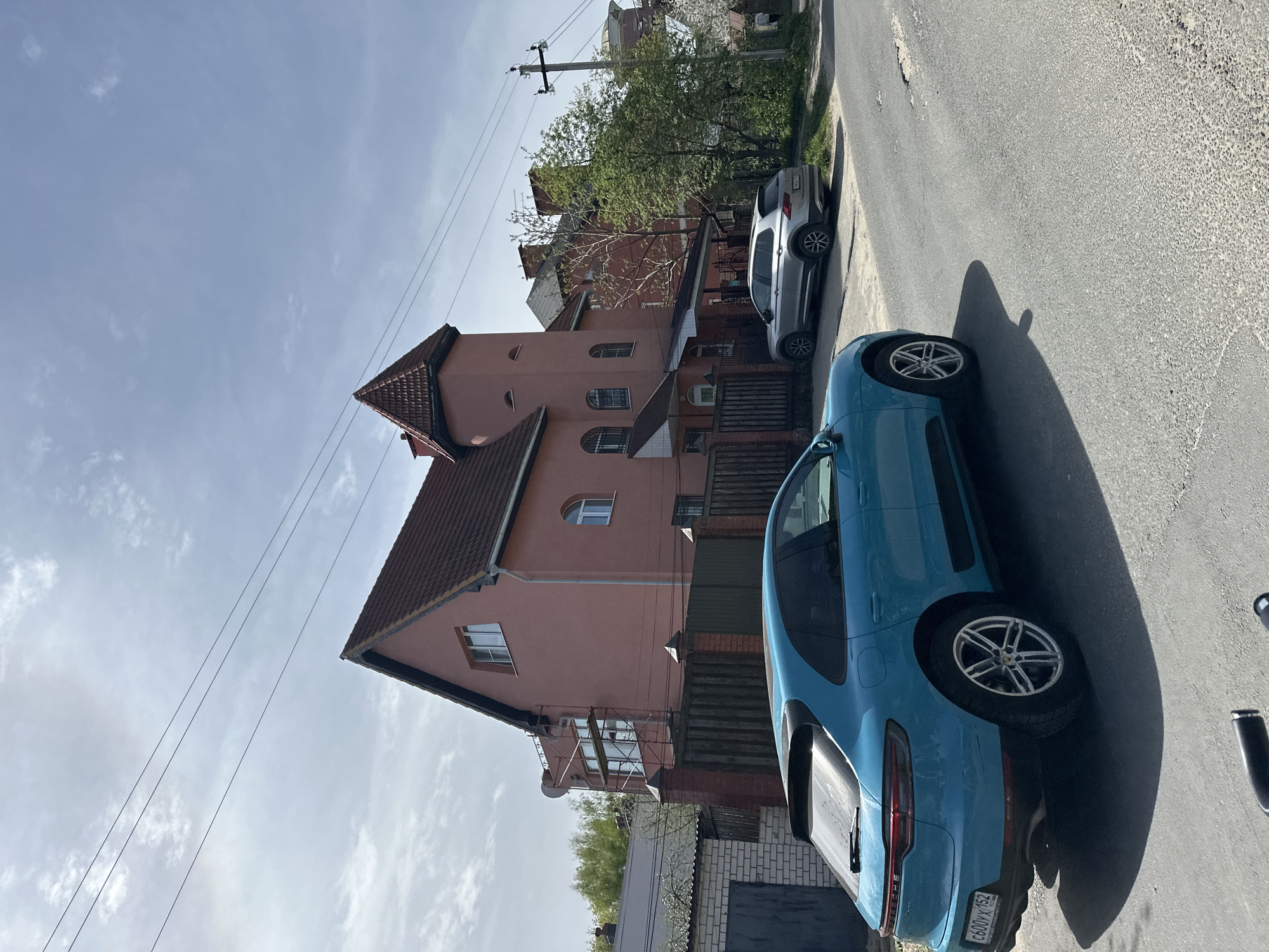 The image shows a street scene in what appears to be a residential area of Nizhny Novgorod, Russia. A light blue Porsche Macan is parked on the right side of the street, along with a light gray SUV. A three-story residential building with a red tile roof is visible in the background. The building has a salmon-colored facade with multiple windows and a balcony on the second floor. Power lines are seen overhead. The road is paved. The sky is blue with some clouds.