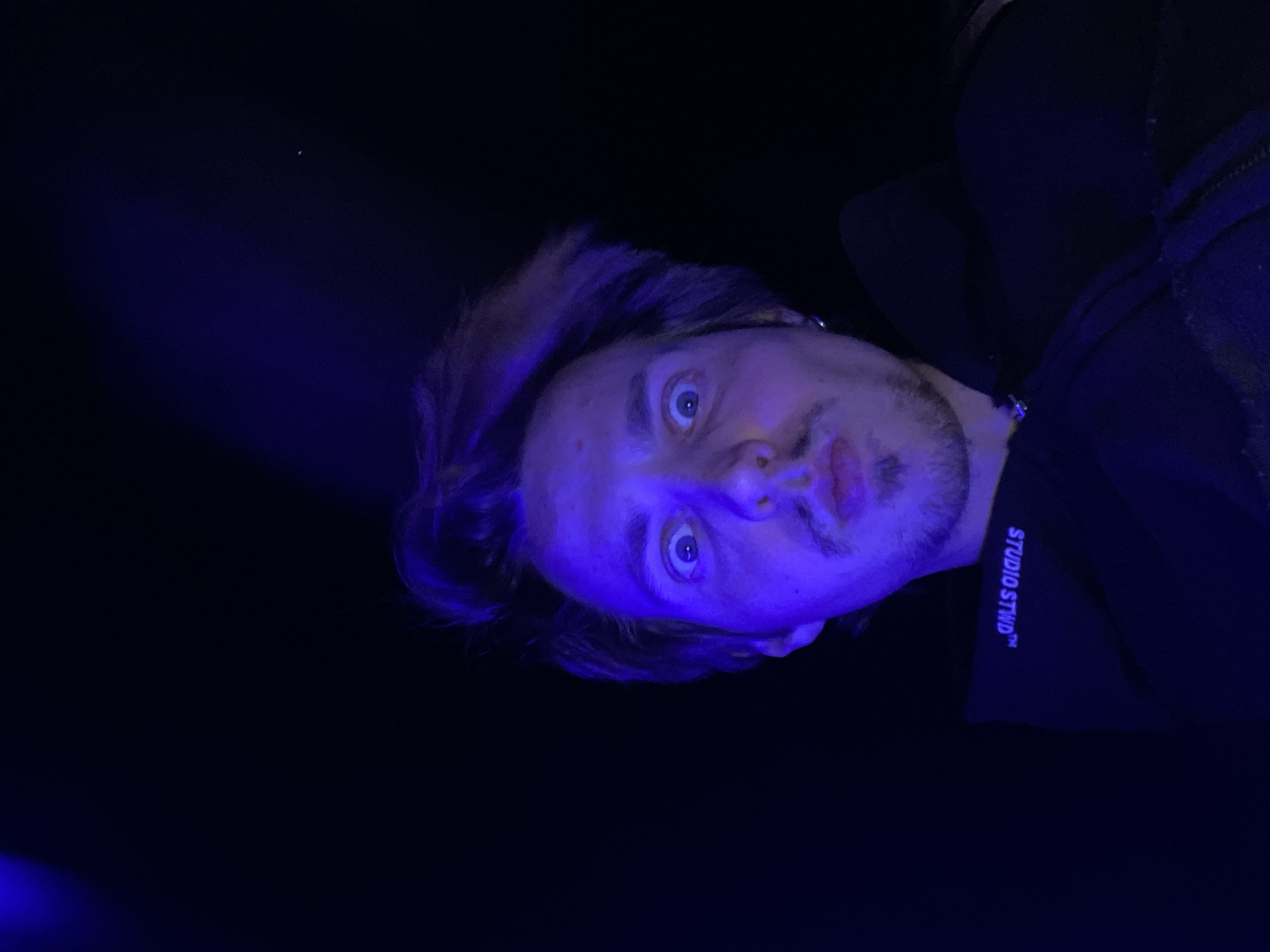 A portrait of a man in a dark environment with blue lighting. The man has light brown hair, a beard, and a surprised expression. He is wearing a dark garment with the words "STUDIO STWD" visible. The location in the city of Bar, Montenegro, is not discernible from the image.