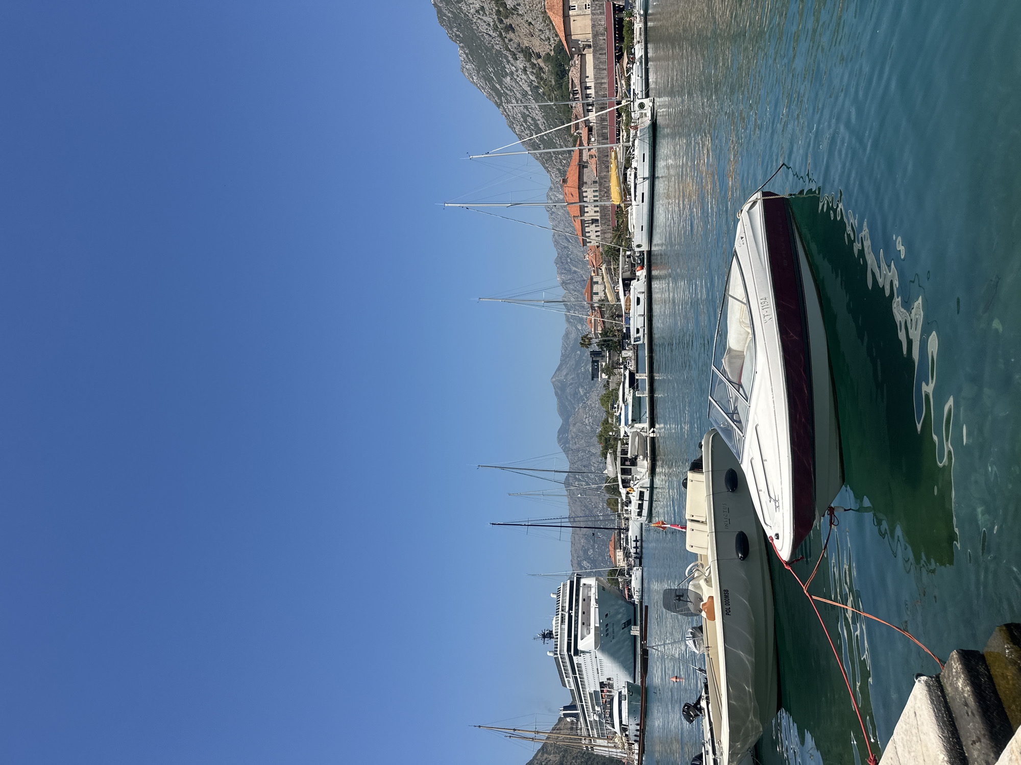 In the city of Kotor, Montenegro, a large cruise ship is docked in a harbor. Several smaller boats and yachts are also moored in the water, with a backdrop of buildings and hills. The sky is clear and blue, and the water is calm and reflects the surroundings. The scene captures a typical day in a picturesque Mediterranean port.