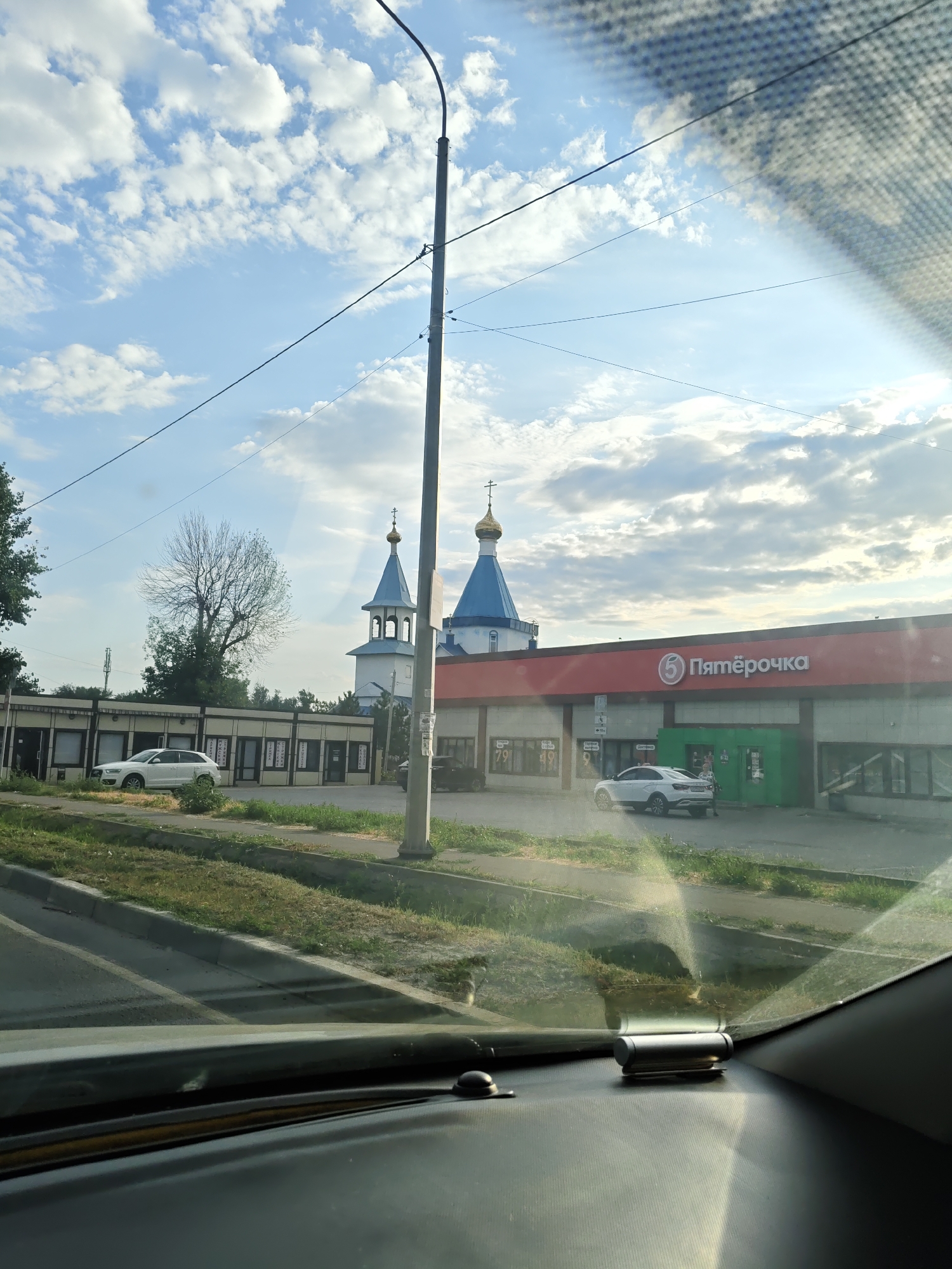 The image depicts a street scene in Novocherkassk, Russia, with a prominent "Pyaterochka" supermarket on the right. A white SUV is parked in front of the supermarket, near a green entrance. To the left of the supermarket, a building with multiple windows is visible, and further left, a church with blue domes and golden crosses stands against a cloudy sky. A street lamp pole is positioned in the foreground. The perspective suggests the photo was taken from inside a vehicle, looking through the windshield, which has some glare. The environment appears to be a suburban or commercial area.