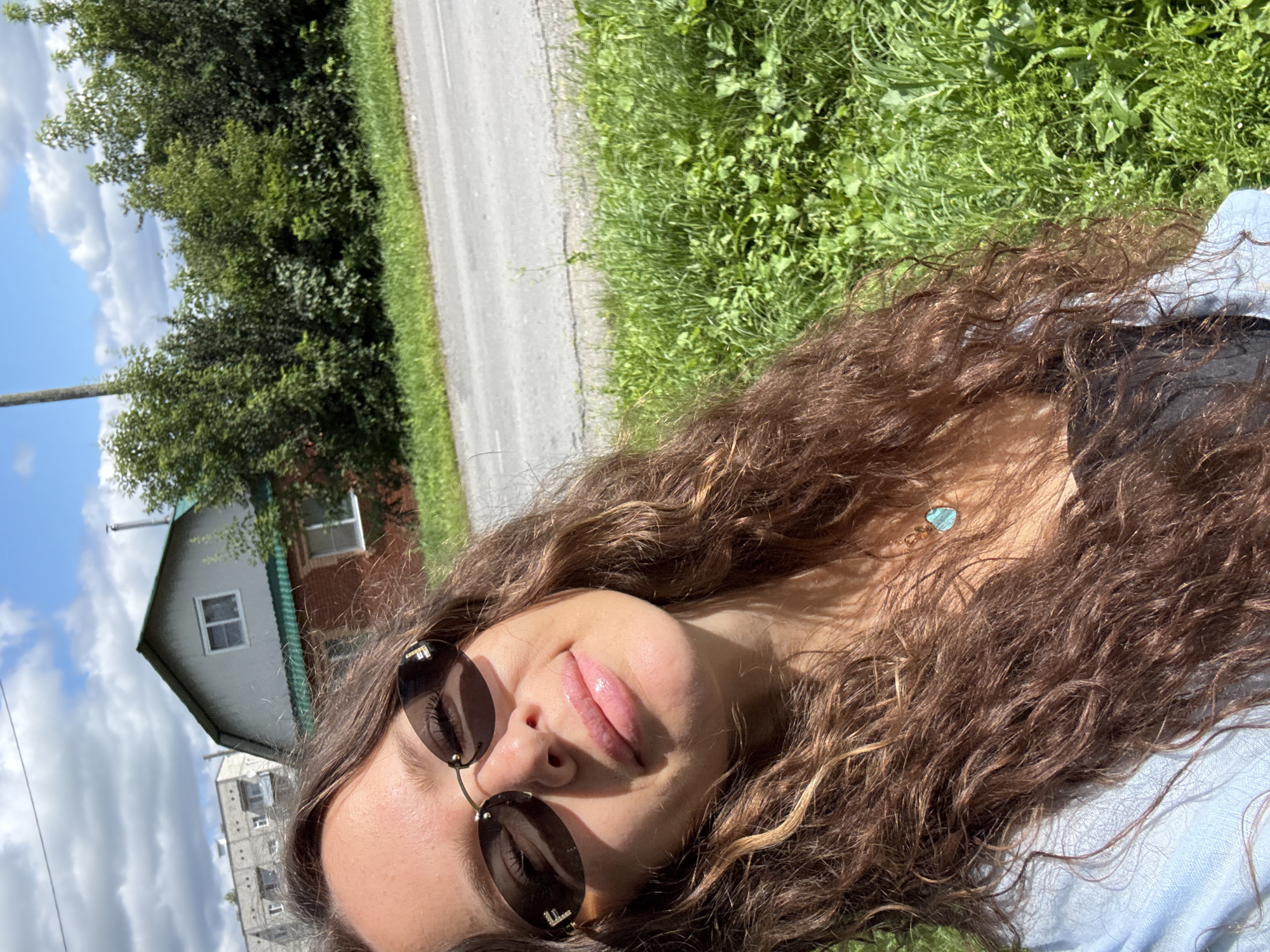 A young woman with long, curly brown hair takes a selfie outdoors on a sunny day. She is wearing oval-shaped sunglasses with gold frames and a light blue shirt. A necklace with a light blue pendant is visible. Behind her, there is a road, a patch of green grass, and trees. A small, light-colored house with a green roof is visible in the background, with what appears to be an apartment building to its left. The sky is blue with scattered white clouds. The scene is likely located in Turinsk, Russia.