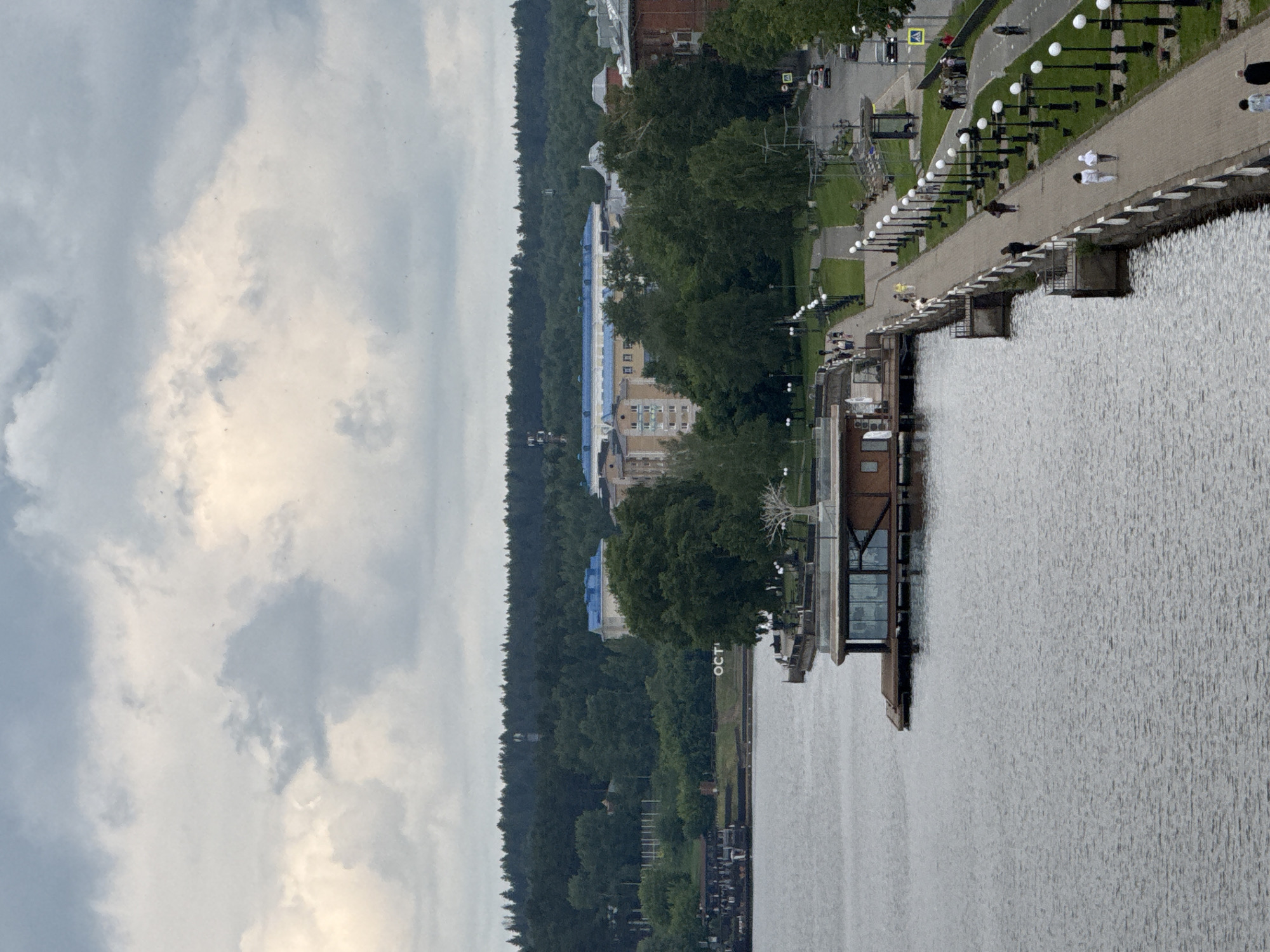 This media file presents a panoramic view of the waterfront in Votkinsk, Russia, captured on a cloudy day. A wide body of water, likely a river or reservoir, fills the foreground and mid-ground, its surface covered in small ripples.
Along the near bank, a paved promenade stretches into the distance, bordered by a low concrete barrier. Numerous dark lampposts, each topped with a distinctive white spherical light, line the walkway. Several people are visible on the promenade; two individuals in light-colored clothing walk together, while another person in dark attire walks separately further down. Extending from the promenade over the water is a modern structure with large windows, possibly a restaurant or viewing platform.
Behind the promenade, a dense line of green trees forms a natural boundary, with a few buildings visible beyond them, including one with a light-colored facade and a blue roof. Further inland and parallel to the promenade, a road can be seen with a yellow pedestrian crossing sign. In the mid-ground, on the grassy bank between the water and the more distant trees, large white letters spelling "ОСТР" (likely part of the Russian word "ОСТРОВ" meaning 'island') are visible. The sky is overcast with heavy grey clouds and brighter patches, indicating diffused daylight. The scene depicts a calm, everyday moment at a public recreational area.