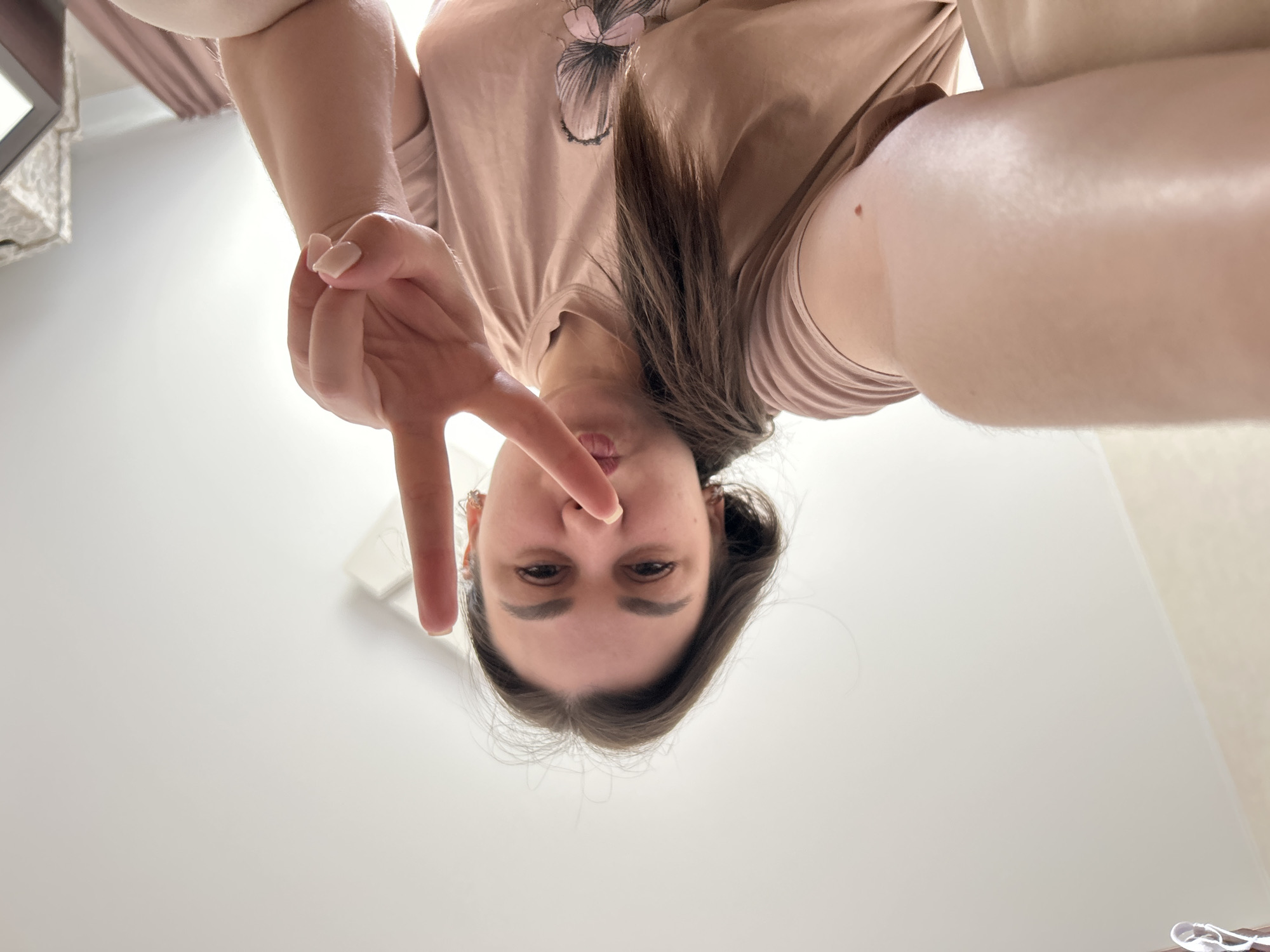 A low-angle selfie photo shows a woman in Chaikovsky, Russia, oriented upside down, looking down at the camera. She is making a "V" sign with her right hand. She is wearing a light brown t-shirt with a hair clip on the front and has long brown hair. The background is a white ceiling and a glimpse of a room with curtains and a shelf.