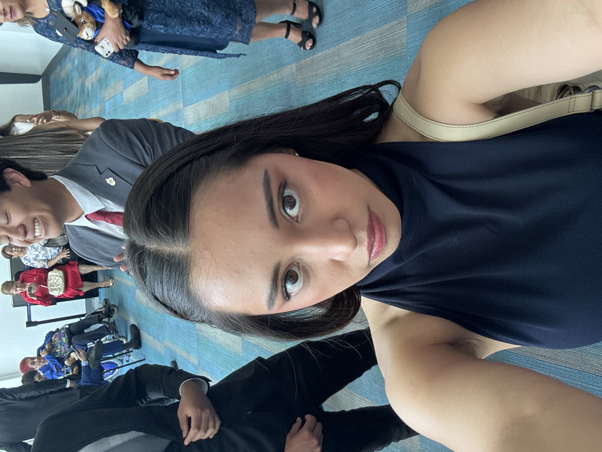 A selfie of a young woman with dark hair and light skin. She is wearing a dark blue top and a beige shoulder bag strap is visible. Behind her, partially visible, are several individuals of varying ages and attire. A man in a gray suit with a red tie is smiling. There's also a woman in a blue lace dress holding a stuffed animal wearing a graduation cap. To the left, people are sitting in wheelchairs and wearing casual clothing. The background appears to be a modern interior with a blue patterned floor. I lack the information to determine whether this location is in Puebla, Mexico.
