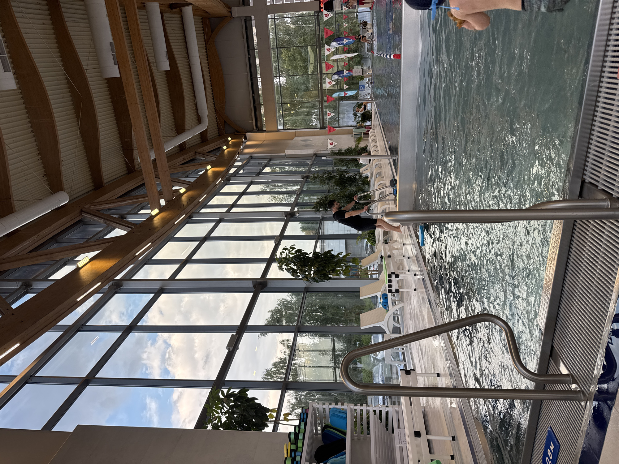 An indoor swimming pool complex with a large glass wall overlooking trees and a cloudy sky is depicted. The pool area features lounge chairs, plants, and a shallow wading area with a ramp leading into it. Several people are present, including a man in a black t-shirt near the ramp, a child in the pool, and other individuals in the background. A shelf filled with swimming accessories like kickboards and goggles is visible on the left. The architecture includes exposed wooden beams and corrugated ceiling panels. The scene suggests a recreational or training facility.