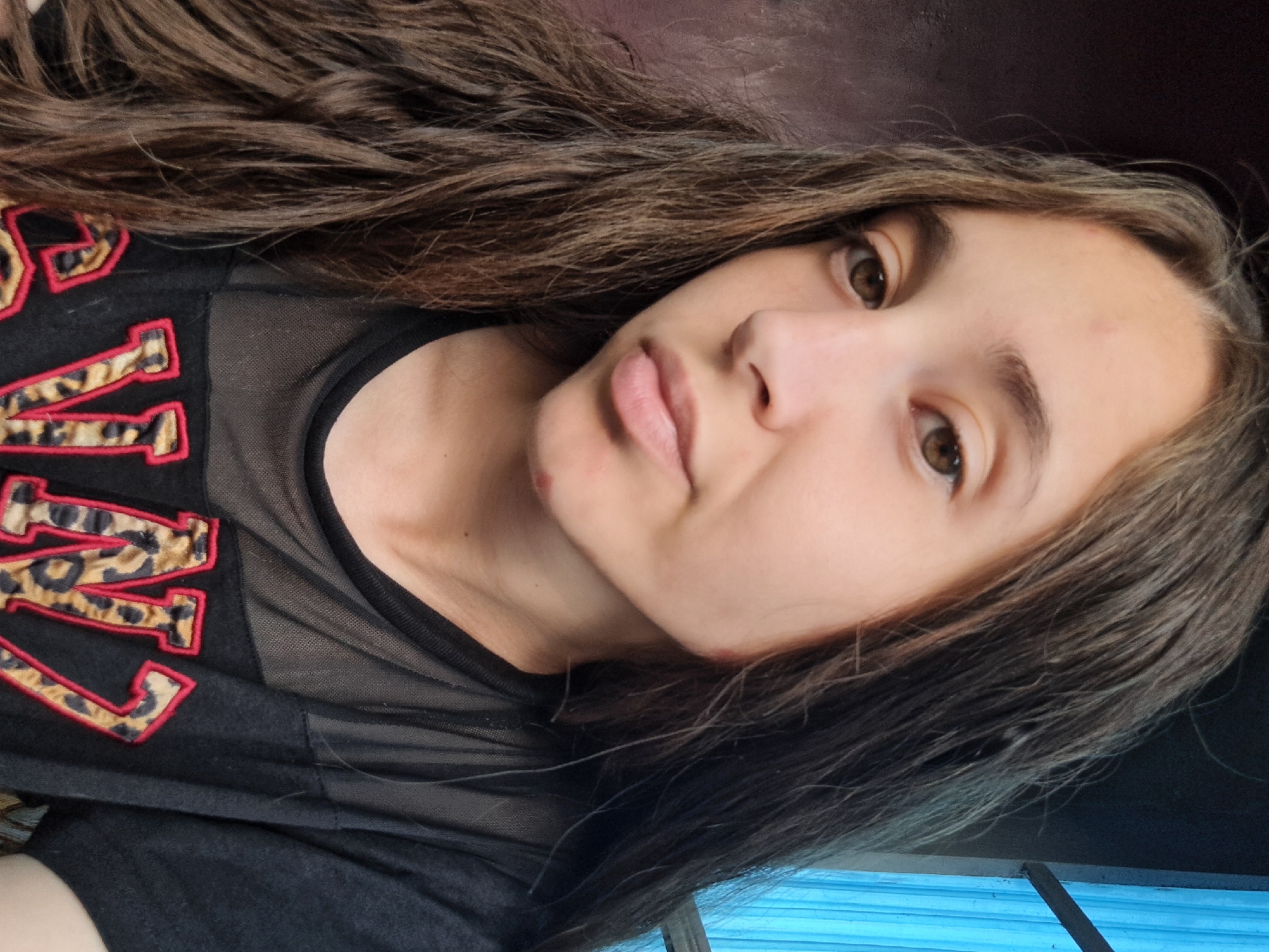 A young female with long, dark brown hair is shown in a close-up portrait. She is wearing a black t-shirt with sheer mesh fabric across the chest and down the sleeves. The t-shirt has large, stylized letters with a leopard print design and red trim. Her skin appears to have some minor blemishes on her forehead and chin. The background on the left is a dark, textured surface, possibly a wall. On the right, a blue, corrugated surface and a black horizontal bar suggest a window or a structure with a blue covering. The lighting suggests an indoor setting, likely a room in Vypasne, Ukraine.
