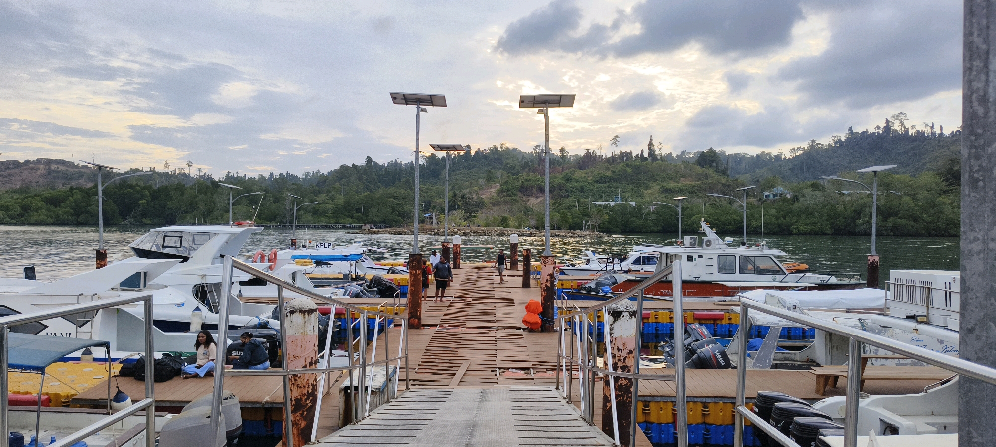 The image shows a dock with several boats moored in the city of Waisai, Indonesia. The sky is cloudy, and the sun is setting, casting a warm glow over the scene. The dock is made of wood and has several solar-powered lights. On the dock, there are several people, some walking and others sitting. The boats are of various sizes, and some appear to be passenger boats, while others may be for fishing or other purposes. In the background, there is a lush green landscape with trees and hills. The water is calm and reflects the sky and the surrounding environment. The overall atmosphere of the image is peaceful and serene.