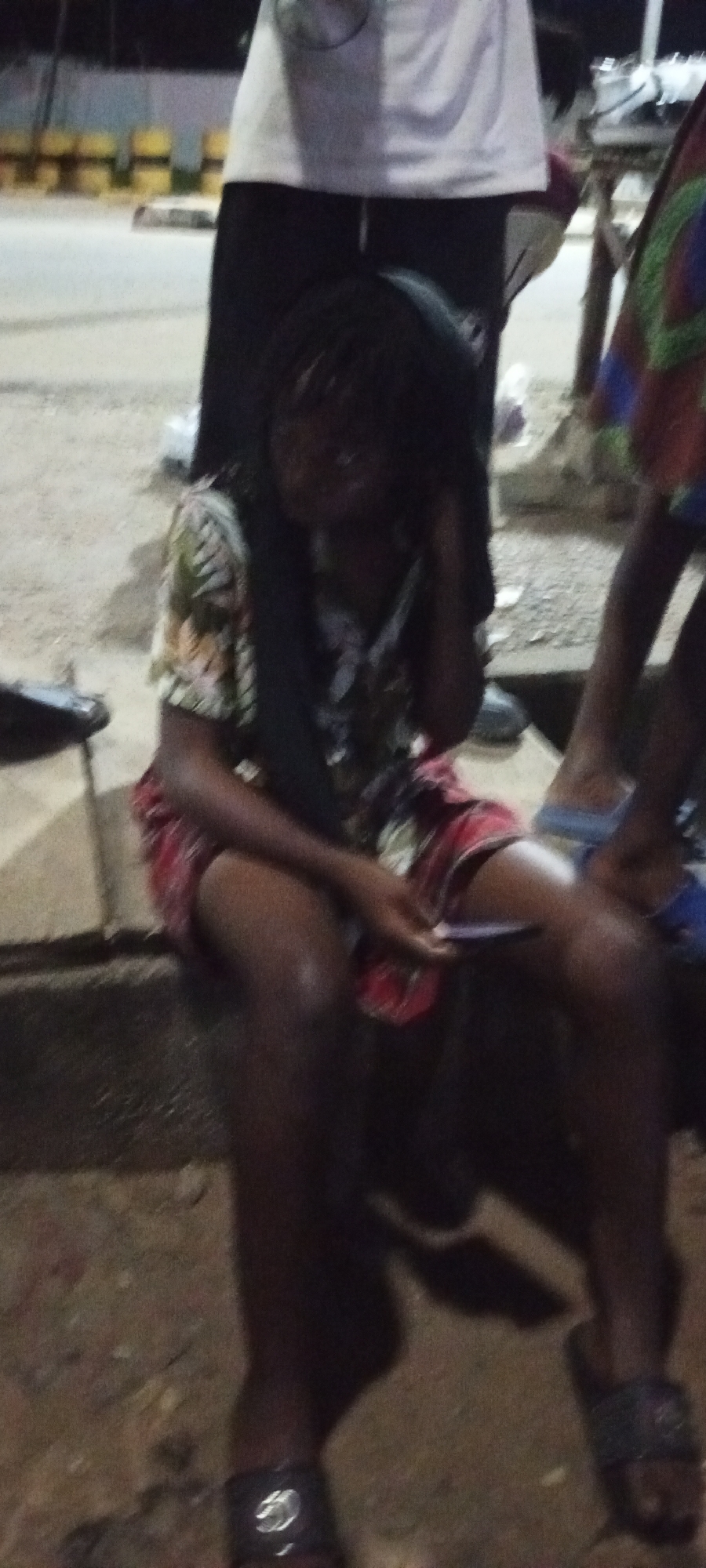 In Adankolo, Nigeria, a young person with dark skin and braided hair is seated on a curb outdoors at night. They are wearing a patterned short-sleeved shirt and a red and white patterned skirt. Their left hand is on their head, and their right hand is holding a cell phone. To their left, a dark object is visible on the ground, possibly a shoe. To their right, another person's legs are visible, wearing blue sandals. The background shows a street with yellow and black striped barriers and other people standing. The scene appears to be a public space, possibly a roadside.