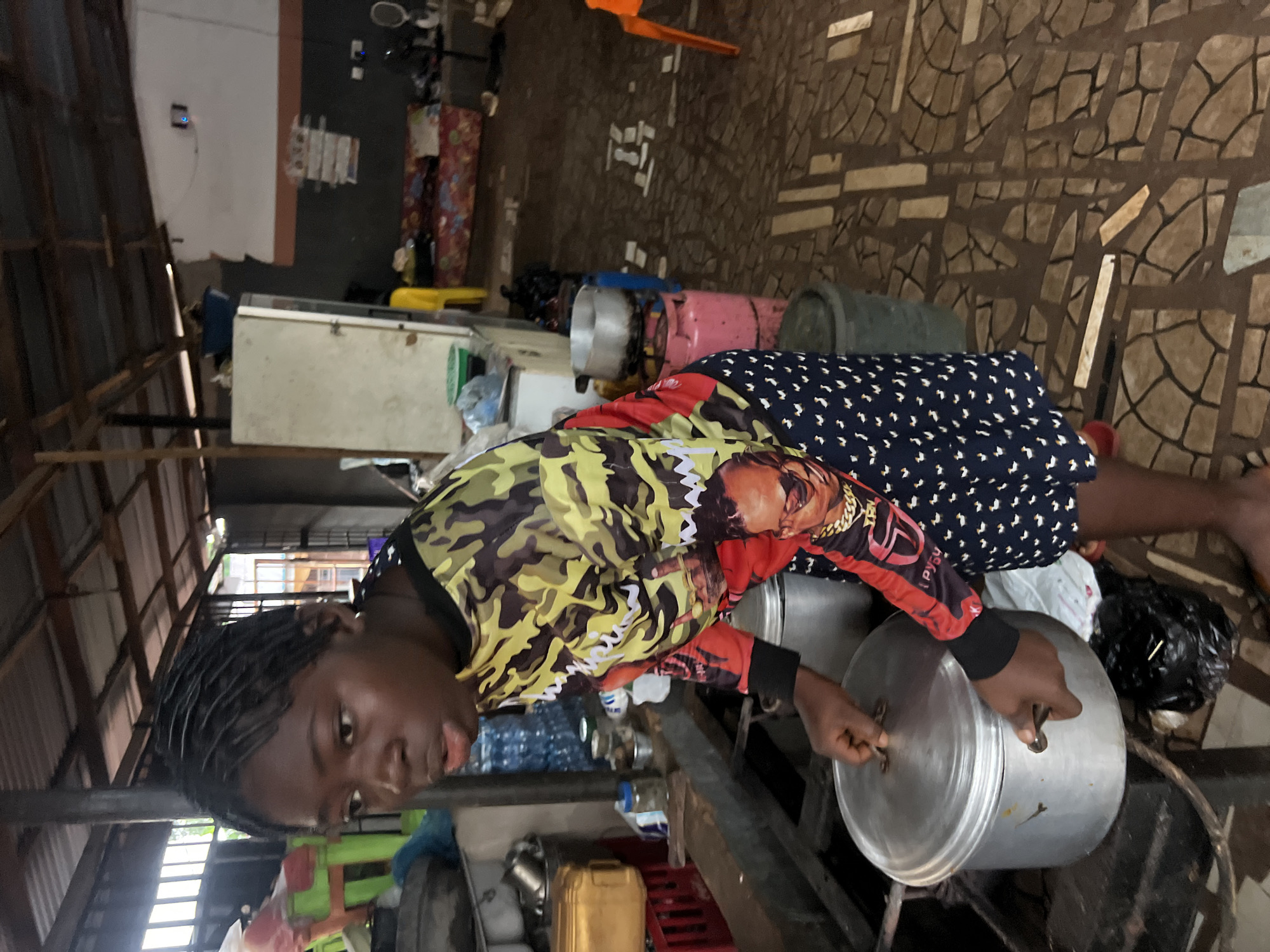 A young Black female with braided hair is standing in what appears to be a marketplace or an open-air kitchen in Nkpor, Nigeria. She is wearing a long-sleeved shirt with a camouflage pattern on the sleeves and upper torso, and a black skirt with a white duck pattern. Her expression is one of concentration as she uses her hands to lift a large silver pot lid.
The environment is rustic and appears to be under a corrugated metal roof supported by wooden beams. In the background, there are various objects commonly found in such a setting: a white refrigerator, a pink gas cylinder, multiple water bottles, metal bowls, a yellow plastic container, and what looks like a large trash bag on the floor. The floor is covered with mosaic-like tiles. There are also glimpses of beds with mattresses in the further background, suggesting a living or sleeping area adjacent to the cooking space. The lighting suggests it is daytime, but the space is somewhat dimly lit.