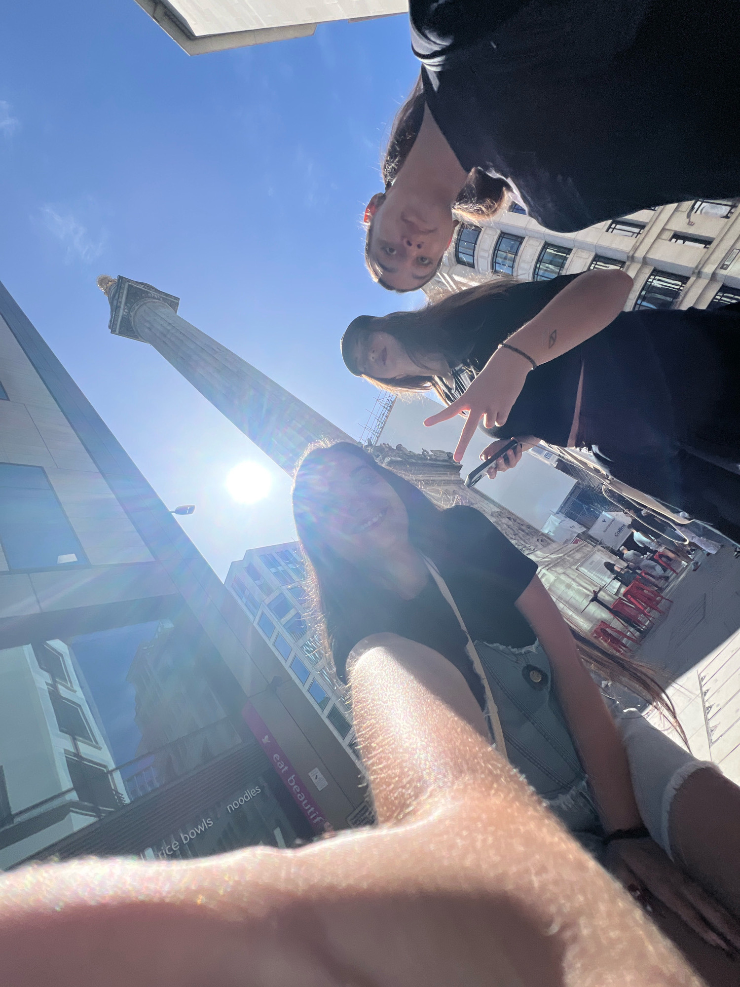 A low-angle, wide shot captures three young women posing for a selfie outdoors on a bright, sunny day in London, United Kingdom. In the background, the monument known as The Monument stands tall against a clear blue sky, partially obscured by lens flare from the sun. The women are positioned in the foreground, with the camera angled upwards. The woman closest to the camera is on the right, with her arm and shoulder in the foreground. She is smiling and her face is slightly blurred. To her left, another woman is also visible, holding up two fingers in a peace sign and holding a smartphone. The third woman is further back and to the left, looking towards the camera. Modern buildings with glass windows line the street, reflecting the surrounding environment. A sign with the text "eat beautiful" and "noodles" is visible on a building to the lower right. The overall scene suggests a tourist outing in a bustling urban setting.