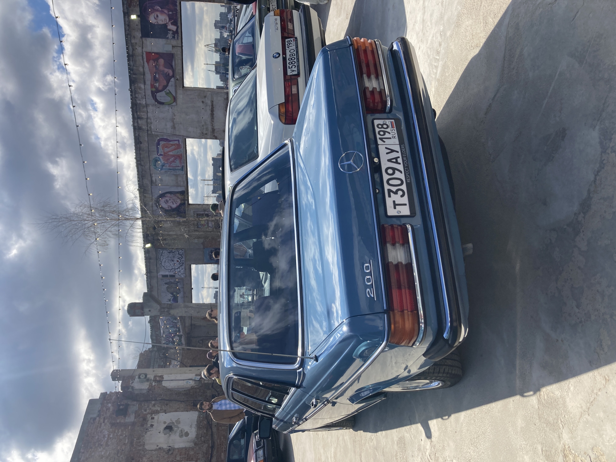 This image depicts an outdoor gathering, likely a car meet, in Saint Petersburg, Russia. The scene is dominated by the rear view of a glossy blue vintage Mercedes-Benz W123 sedan, which has "200" emblazoned on its trunk. Its Russian license plate reads "T309AY 198 RUS", with an additional phrase "RESPECT YOUR ELDERS" printed on a strip below the plate. Parked behind it, a white BMW E34 sedan is partially visible, bearing the Russian license plate "У588ВО 198 RUS". Both vehicles are parked on a concrete surface that shows distinct shadows from the bright sunlight.
The backdrop consists of an old, weathered brick building, possibly an industrial structure repurposed for events. Its walls feature various murals, graffiti, and embedded mirror-like panels that reflect the cloudy sky and distant cityscape, including what appear to be cranes. Strung across the scene are decorative string lights, adding to the event atmosphere.
Several people are scattered throughout the area, casually observing the cars or socializing. Their reflections are visible in the car windows and the mirrored building panels. The time of day appears to be daytime, with a mix of sun and clouds creating varied lighting conditions. The overall impression is one of a relaxed social event focused on classic cars within an urban, industrial-chic setting.