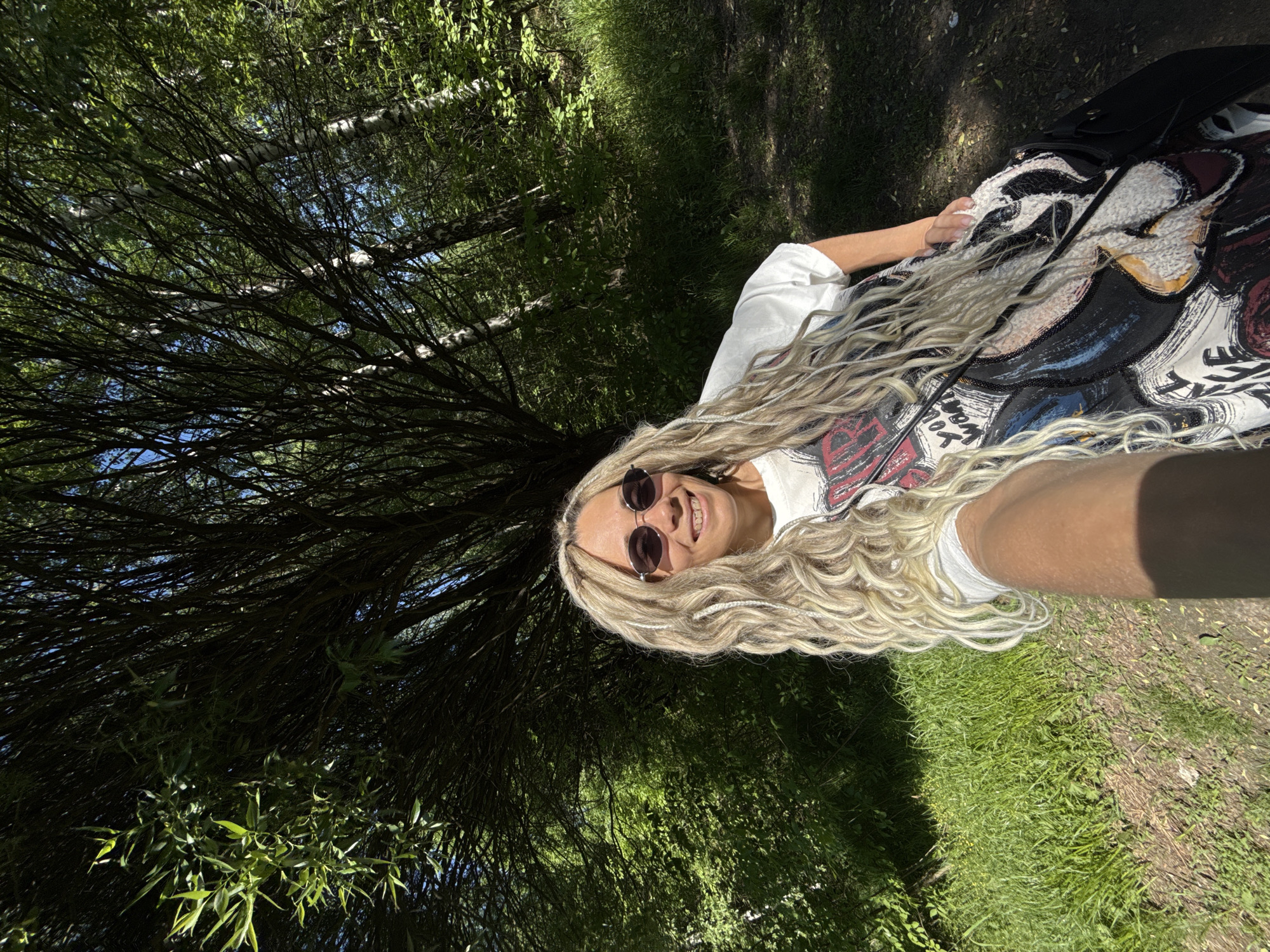 A medium shot depicts a woman with fair skin and long, blonde, wavy hair, wearing sunglasses and a white t-shirt with a graphic design. She appears to be in an outdoor, wooded area. The setting includes foliage, green grass, and branches overhead, suggesting a forest or park. The scene is likely in Rusanovo, Russia.