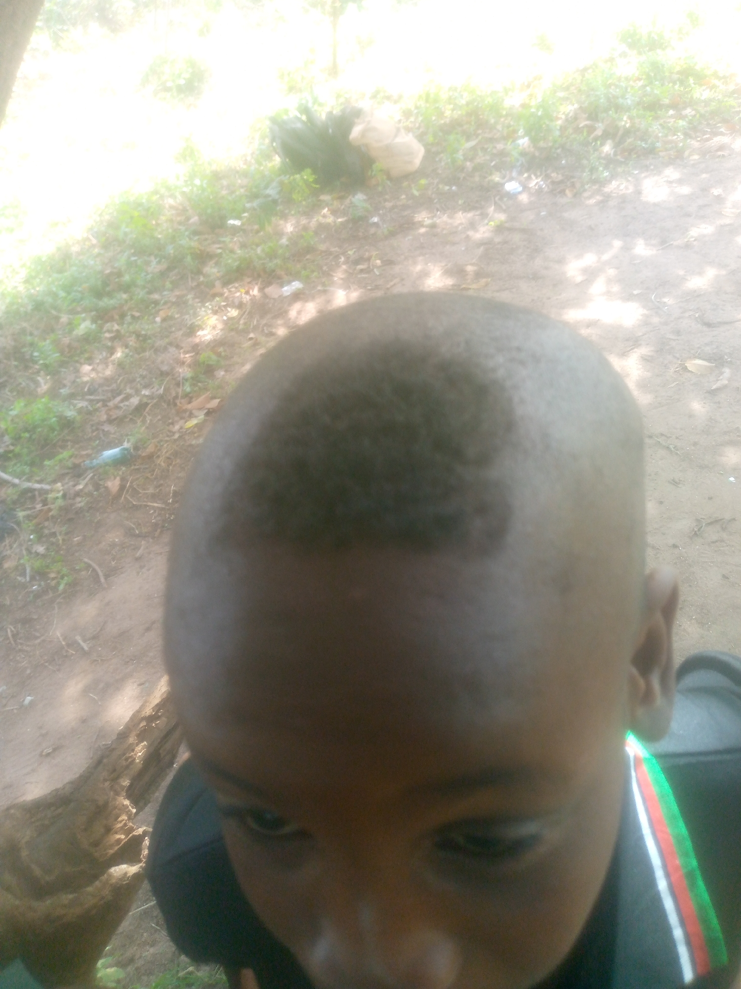 A close-up, high-angle shot shows a young Black person with a recently cut hairstyle featuring a defined, square section on the top of their head. The person is wearing a dark-colored shirt with a trim featuring green, white, red, and black stripes near the neckline. The background shows dappled sunlight, dirt, foliage, and trash. The location in the city of Unknown, Unknown cannot be determined from the image.