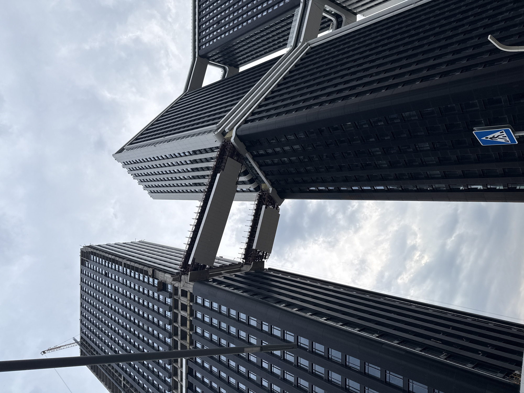 The image captures a low-angle view of two modern, high-rise buildings under a cloudy sky. The buildings are connected by bridge-like structures. The buildings have a dark exterior with repetitive window patterns. A blue and white pedestrian crossing sign is visible on one of the buildings. A construction crane can be seen on the left side of the frame. The location is Moscow, Russia.