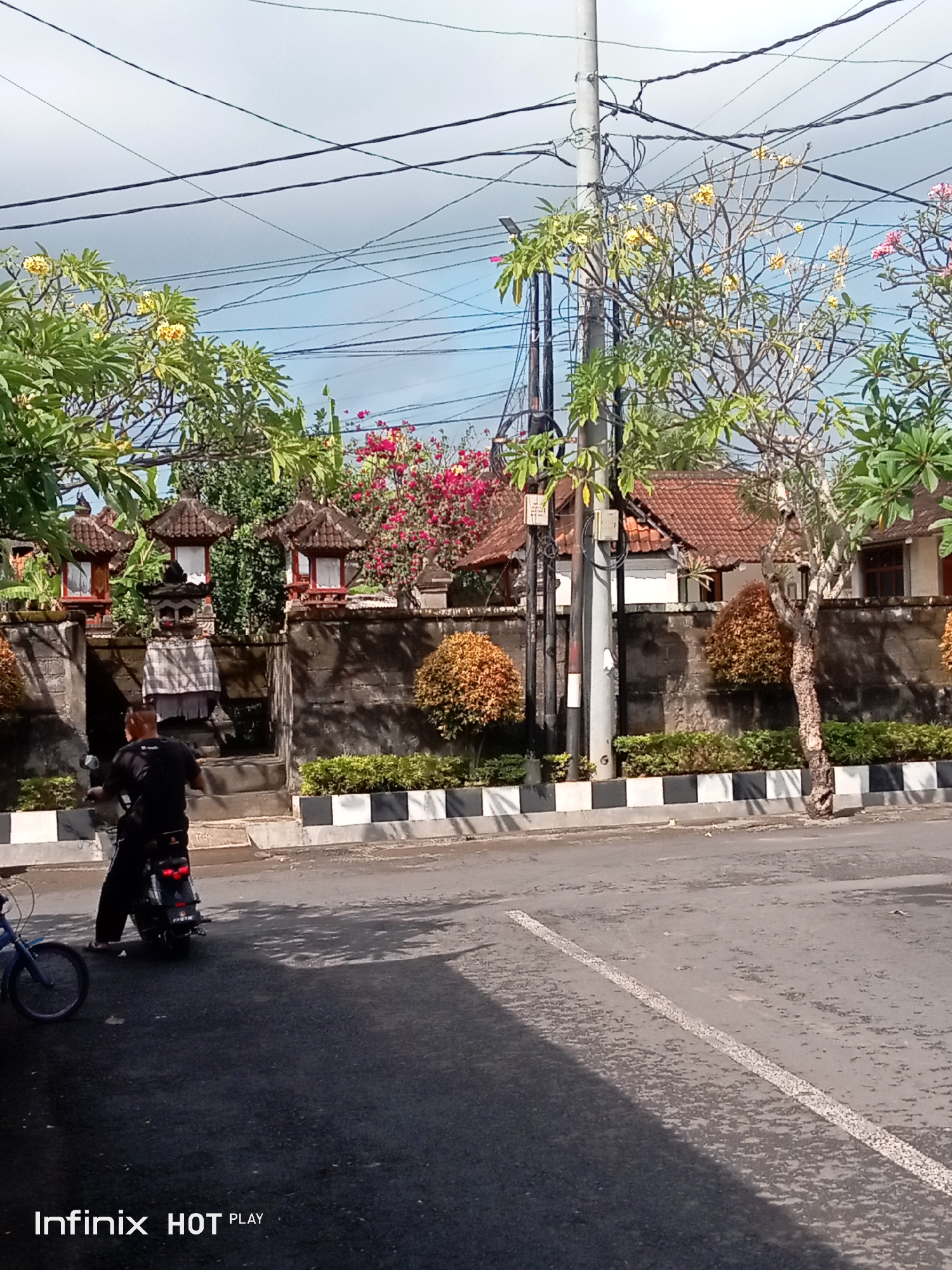 The image shows a street scene in Blahbatuh, Indonesia, with a person riding a scooter. The scooter is dark colored and the rider is wearing a black t-shirt and dark pants. The scooter is moving away from the camera. In the background, there is a stone wall with several decorative stone lanterns. Beyond the wall, there are buildings with red tile roofs and lush greenery, including flowering trees with yellow and pink blossoms. A utility pole with numerous wires is also visible. The road is paved and has white lines marking the lanes. A blue bicycle is parked on the left side of the image. The sky is partly cloudy. The overall impression is of a typical street in a tropical urban environment.