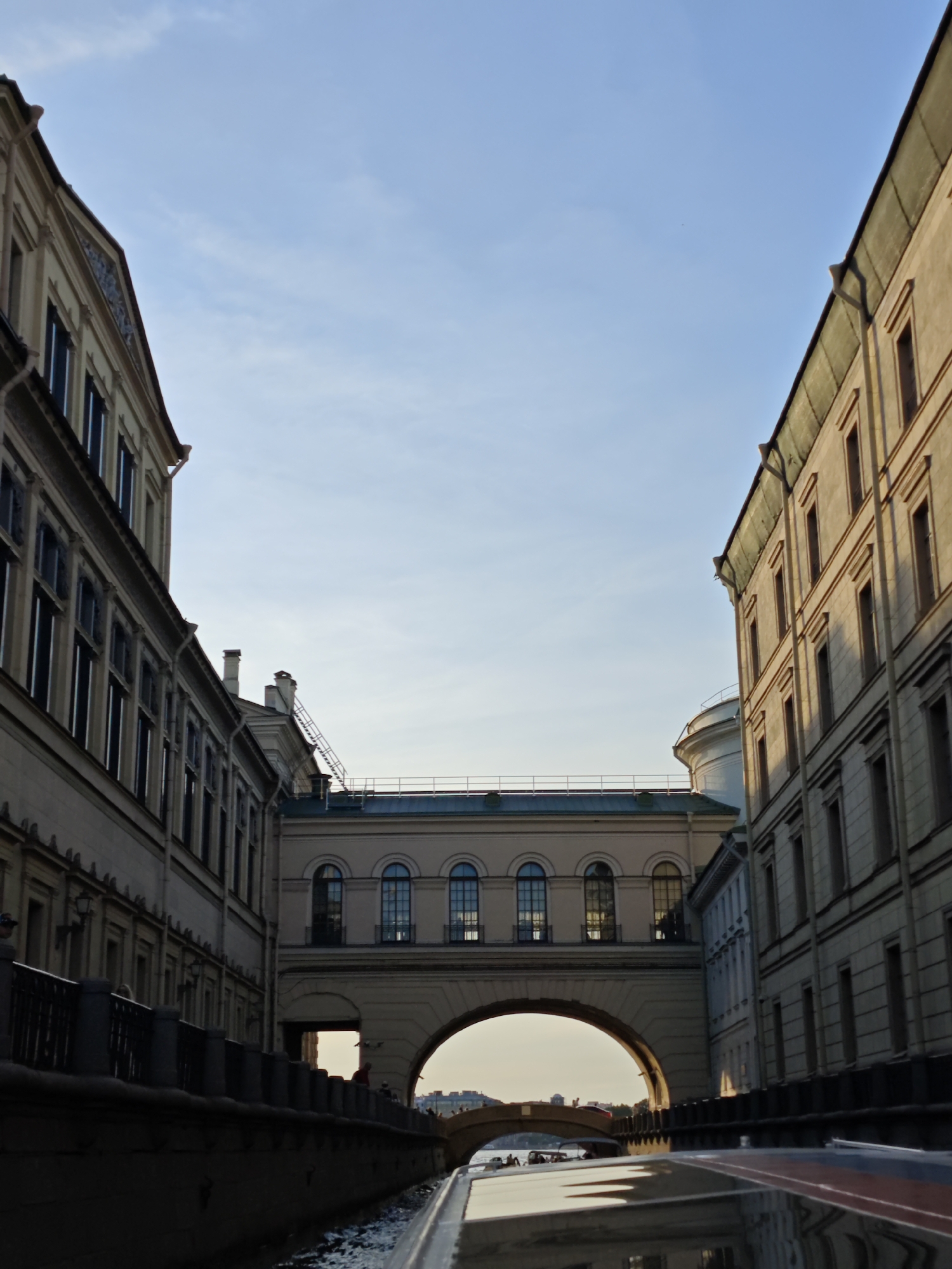 The image shows a view from a boat traveling along a canal, with buildings on either side. The canal is bordered by a dark stone embankment with a black metal railing. In the foreground, the glossy surface of the boat reflects the sky and buildings.
Dominating the scene is a large, arched structure that bridges the canal, connecting two buildings. This bridge-like structure has a light-colored facade with a row of arched windows. Above and behind this structure, another building is visible with a curved, white dome or turret.
On the left and right sides of the canal, tall, multi-story buildings line the banks. These buildings have a light beige facade and a dark green roof. The windows on these buildings are tall and narrow. The building on the left appears to be older, with visible architectural details like pilasters and decorative elements above the windows. The building on the right is more uniform in its window placement.
In the distance, seen through the arch of the connecting structure, is a hint of another bridge and some indistinct structures. The sky is clear and blue, with some wispy clouds. The lighting suggests it is late afternoon or early evening, with the sun casting long shadows. There are a few people visible on the embankment and a small boat or two in the distance, suggesting this is a public area or a tourist waterway.
