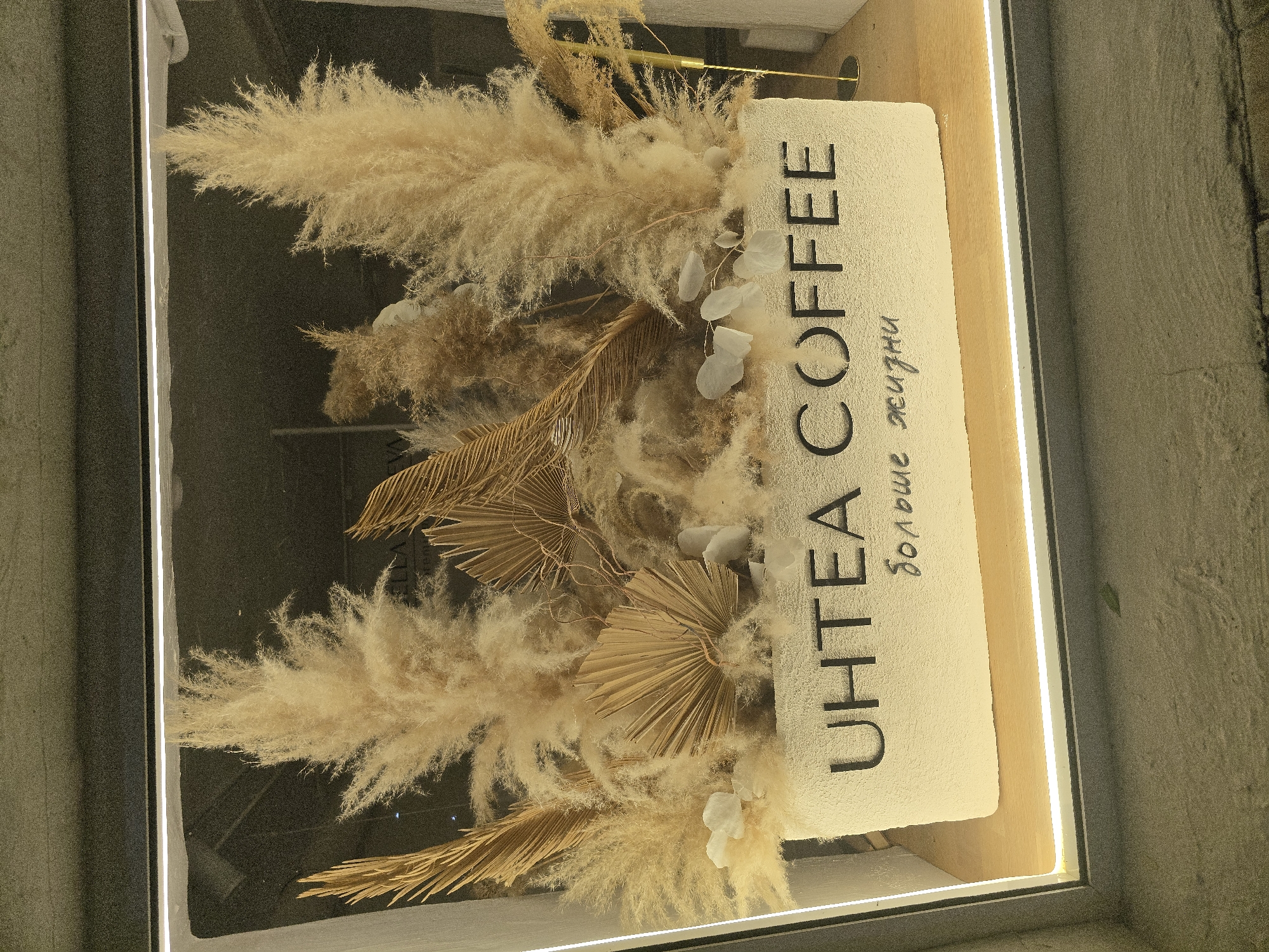The image shows a storefront sign for "UHTEA COFFEE" located in an unidentified city. The sign is predominantly beige with black lettering and features decorative dried flowers and leaves. The environment appears to be a modern urban setting.