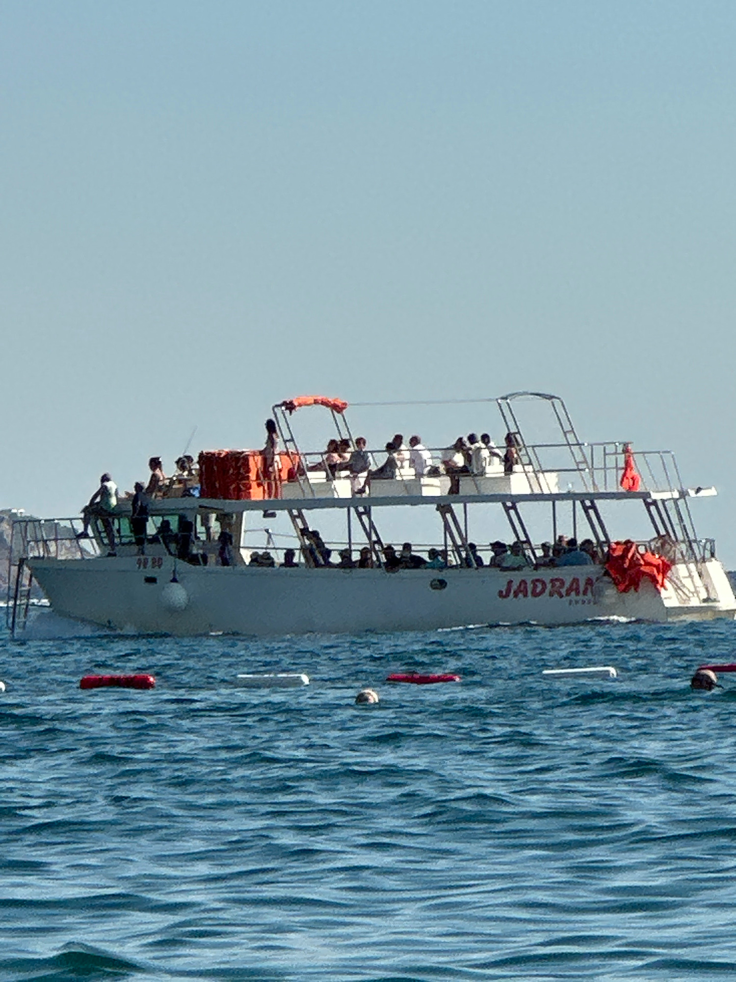 The image shows a boat named "JADRAN" with numerous passengers on board, cruising on a body of water. The boat has two levels, with people visible on both the lower and upper decks. Several individuals are standing and sitting, some appearing to be enjoying the ride. Red life vests are visible on the back of the boat. In the foreground, the blue water exhibits small waves, and several buoys, some red and some white, are floating on the surface. The sky is clear and blue, indicating a sunny day. The location is identified as the city of Boreti, Montenegro.