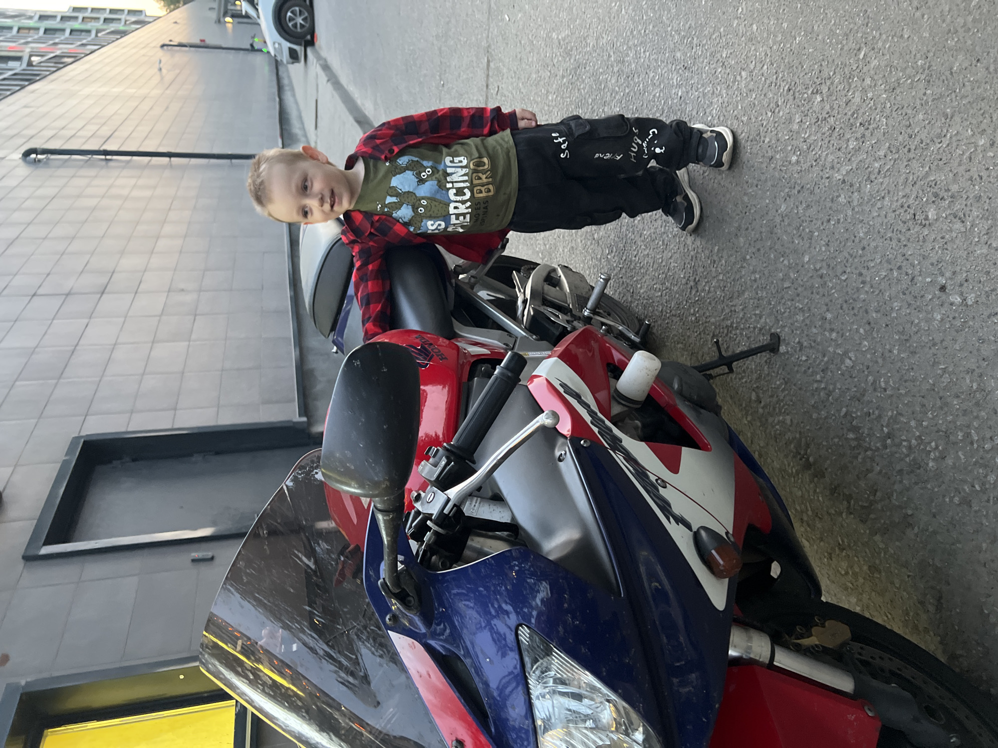 A young boy is standing next to a red, white, and blue Honda motorcycle on a paved surface. He wears a red plaid shirt over a green t-shirt, black pants with writing on them, and sneakers. The motorcycle is parked near a gray building with yellow accents. The location is identified as the city of Mytishchi, Russia.