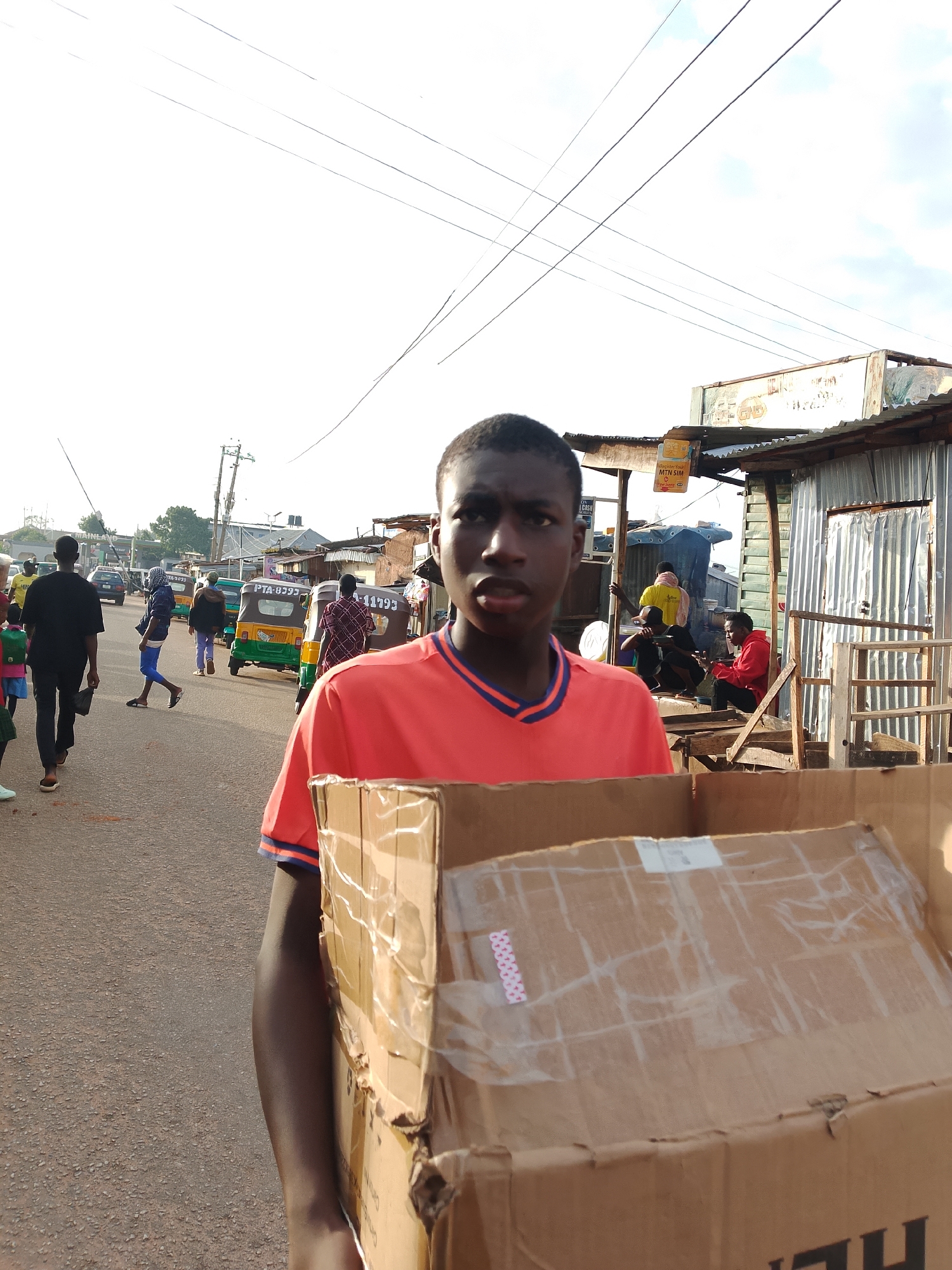 In Jos, Nigeria, a young boy in a bright orange shirt with blue trim walks towards the camera, carrying a large cardboard box. The box is taped shut with clear packing tape, and a pink, digital-looking sticker is visible on its surface. The boy's expression is neutral, and his gaze is directed forward.
Behind him, the street is bustling with activity. A variety of people are walking and moving about. Several motorized tricycles, known locally as "keke napeps", are parked and in motion. One green and yellow keke napep has "PTA-0273" visible on its side. Another brown keke napep displays "11793". In the background, small shops with corrugated metal siding line the street. The sky is overcast, with power lines crisscrossing above. The road surface is asphalt, showing signs of wear. Further down the street, cars and more people are visible, suggesting a busy urban environment.