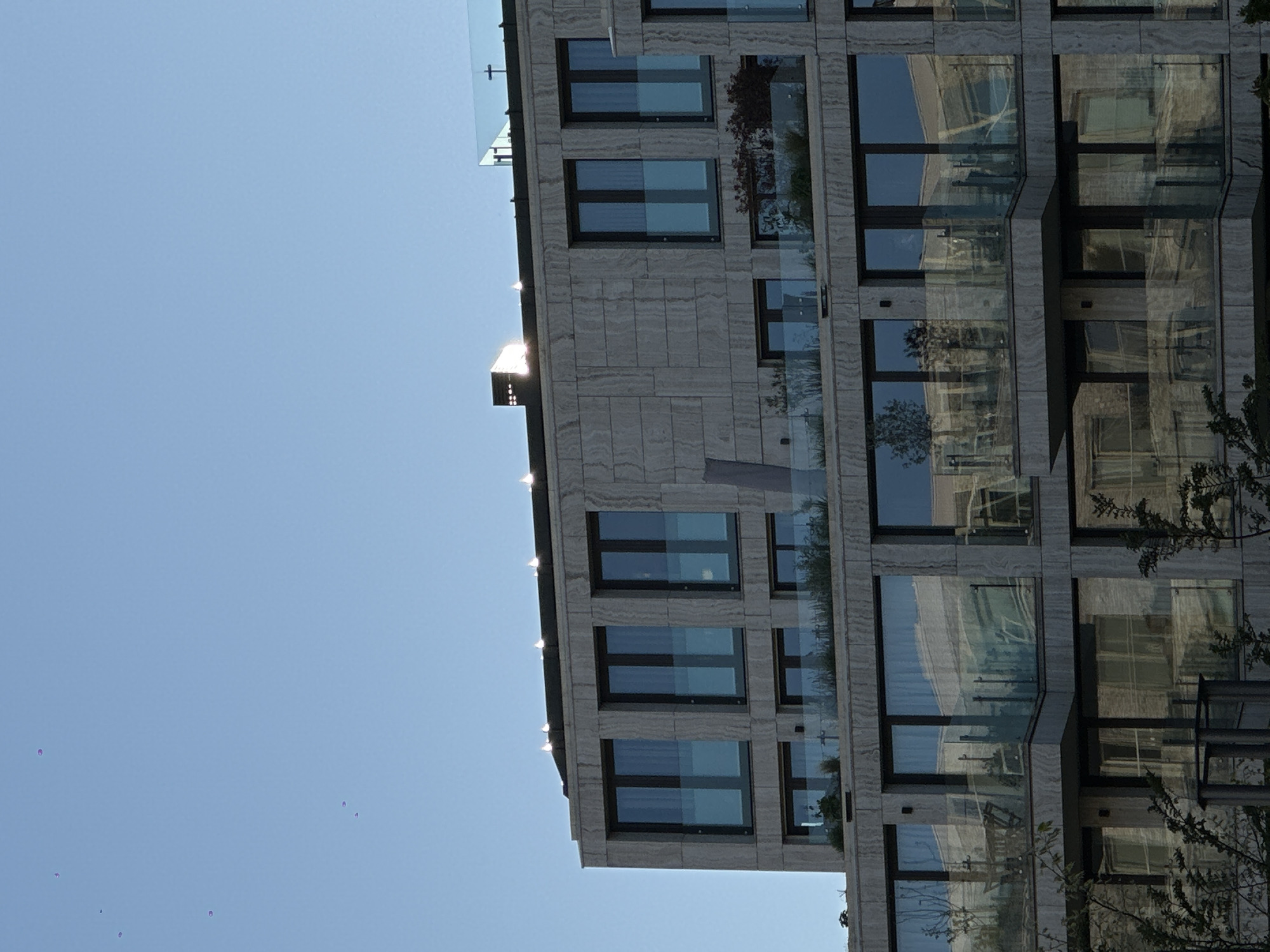 Here's a detailed description of the image:
**Scene:** An architectural view of a modern building, likely a residential complex. The building features a grid-like facade with multiple windows and glass-paneled balconies. The facade is clad in a light-colored material with a textured, possibly stone-like, finish. Sunlight reflects off the building, creating bright highlights along the edges. The windows appear dark, reflecting the sky and surrounding environment. There are green plants growing on some of the balconies.
The sky is clear and blue.
**Location:** Although not definitively stated, based on the architectural style (modern and clean lines), and the presence of balconies with greenery, it is plausible to be Amsterdam, Netherlands, a location known for modern buildings.