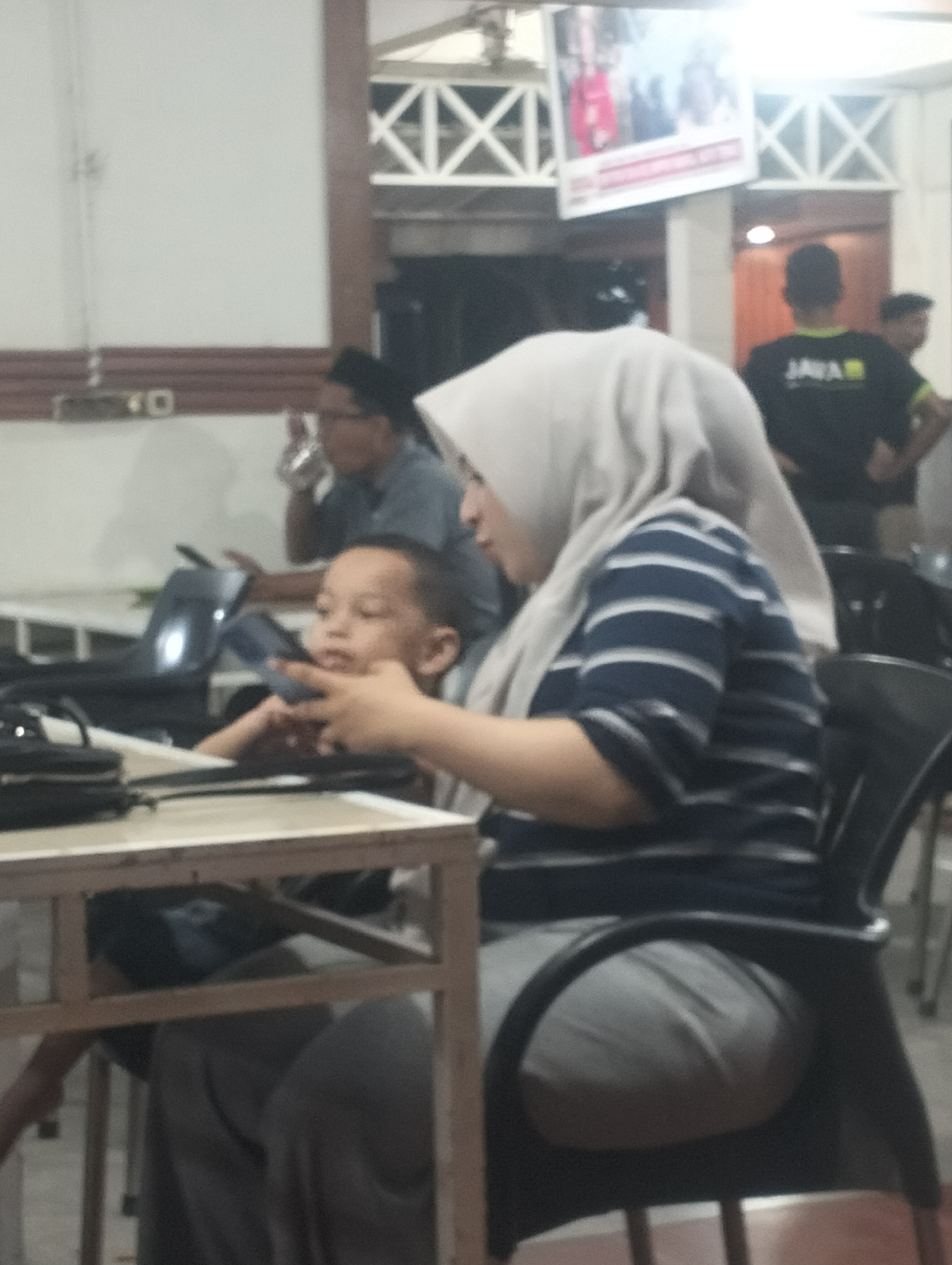 The image depicts a casual indoor gathering, likely in a restaurant or cafe, in Lhokseumawe, Indonesia. In the foreground, a young boy is seated, looking intently at something in front of him, possibly a phone. He is wearing a light-colored shirt. Beside him, a woman in a light grey hijab and a blue and white striped shirt is also seated, seemingly engaged with the boy. The background shows other individuals, including a man in a black t-shirt with the word "JAVA" on it, and another person partially obscured. The environment appears to be a dining area with tables and chairs, and a large screen displaying an image is visible on the wall in the background. The lighting suggests it is either daytime with indoor illumination or evening.