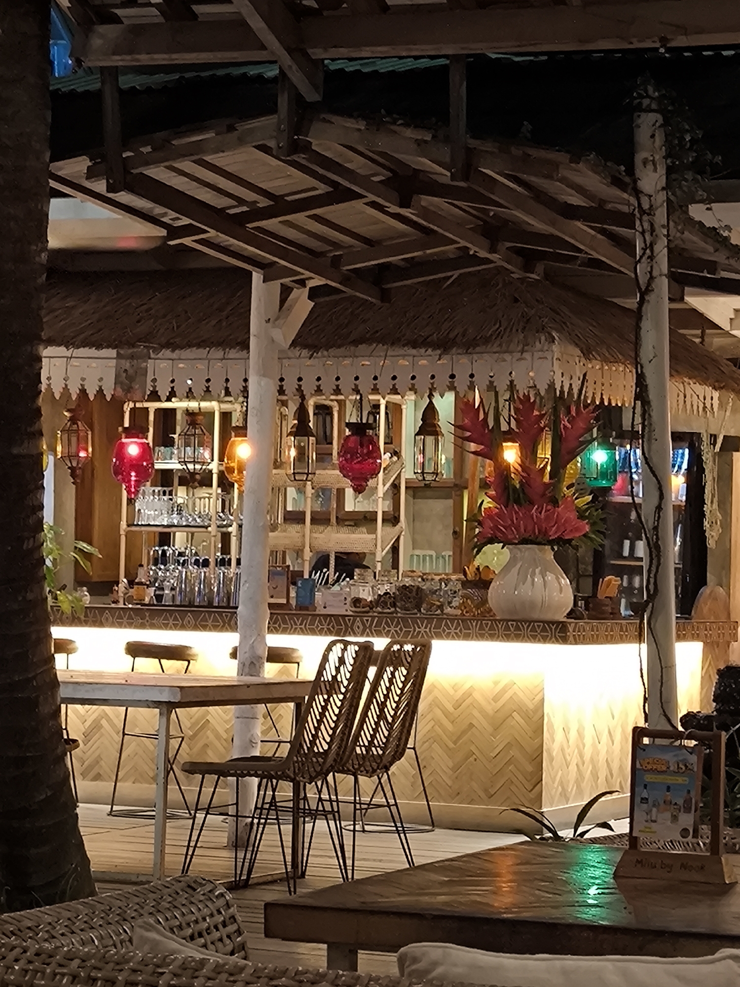 The image depicts a warm and inviting tropical-themed outdoor bar or restaurant at night, set in North Kuta, Indonesia. The scene is illuminated by ambient lighting, creating a relaxed and cozy atmosphere.
The central feature is a prominent bar counter, which glows with a warm light from beneath, highlighting its herringbone-patterned wooden facade. Behind the bar, shelves are neatly stocked with glassware. A large white vase on the counter holds a vibrant arrangement of red and pink tropical flowers, likely ginger or heliconia. Above the bar, several colorful lanterns in red, orange, and green hues hang decoratively, casting soft light.
The structure itself is built with dark wooden beams and features a thatched roof, typical of tropical architecture. On the far left, a section of a palm tree trunk is visible, reinforcing the exotic setting. A white pillar, partially adorned with vines, supports the overhead structure on the right side of the frame.
In the foreground, there are wooden tables and woven rattan chairs, indicating seating for guests. On one of these tables, a small wooden A-frame sign is displayed, with the text "Menu by Nook" clearly visible at its bottom. The upper part of the sign also mentions "VODKA 15%" alongside images of liquor bottles.
The time of day is clearly night, as evidenced by the dark background and the reliance on artificial lighting. The overall ambiance suggests a laid-back and pleasant environment for socializing and enjoying drinks in a tropical locale. No people are visible, allowing the focus to remain on the establishment's charming decor and inviting setup.