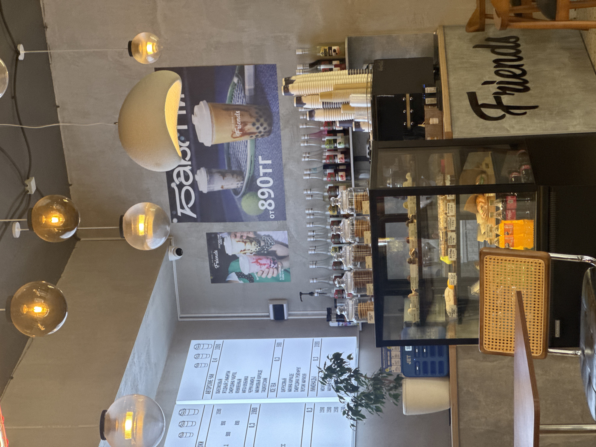 This media file captures an interior view of a cafe with a "Friends" theme, likely located in Kokshetau, Kazakhstan. The scene is set in a modern, casual environment with concrete walls and a minimalist aesthetic. Prominently displayed are several hanging globe lights, casting a warm glow. On the walls, there are posters, one featuring bubble tea with the text "Friends" and "от 890тг", indicating a price point. Another poster displays the word "Friends" in large, stylized script.
The cafe appears to be set up for service, with a display cabinet showcasing desserts and bottled beverages. Behind the counter area, shelves are stocked with various syrups and what appear to be paper cups, suggesting a focus on drinks like bubble tea and other custom beverages. A menu board is visible, listing different types of tea and lemonades with their respective sizes and prices in Russian (e.g., "АВТОРСКИЕ ЧАЙ", "ICE TEA", "ЛИМОНАДЫ").
In the foreground, a cane-backed chair and part of a table are visible, implying seating arrangements for customers. A small plant adds a touch of greenery to the space. The overall impression is that of a contemporary beverage shop, likely operating during daytime hours, judging by the lighting and the active setup. There are no people clearly visible in the image, but the presence of the menu and display suggests it's open for business.